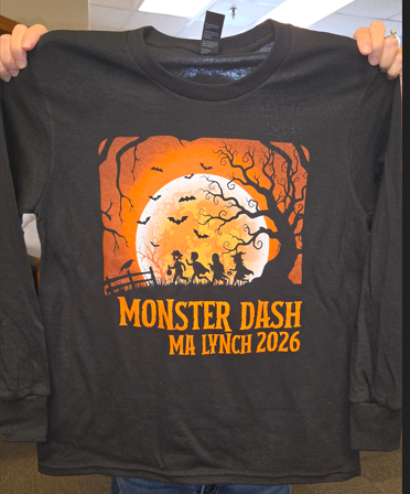 A person holds up a black long-sleeve shirt with an orange Halloween graphic and text that reads "MONSTER DASH MA LYNCH 2026".