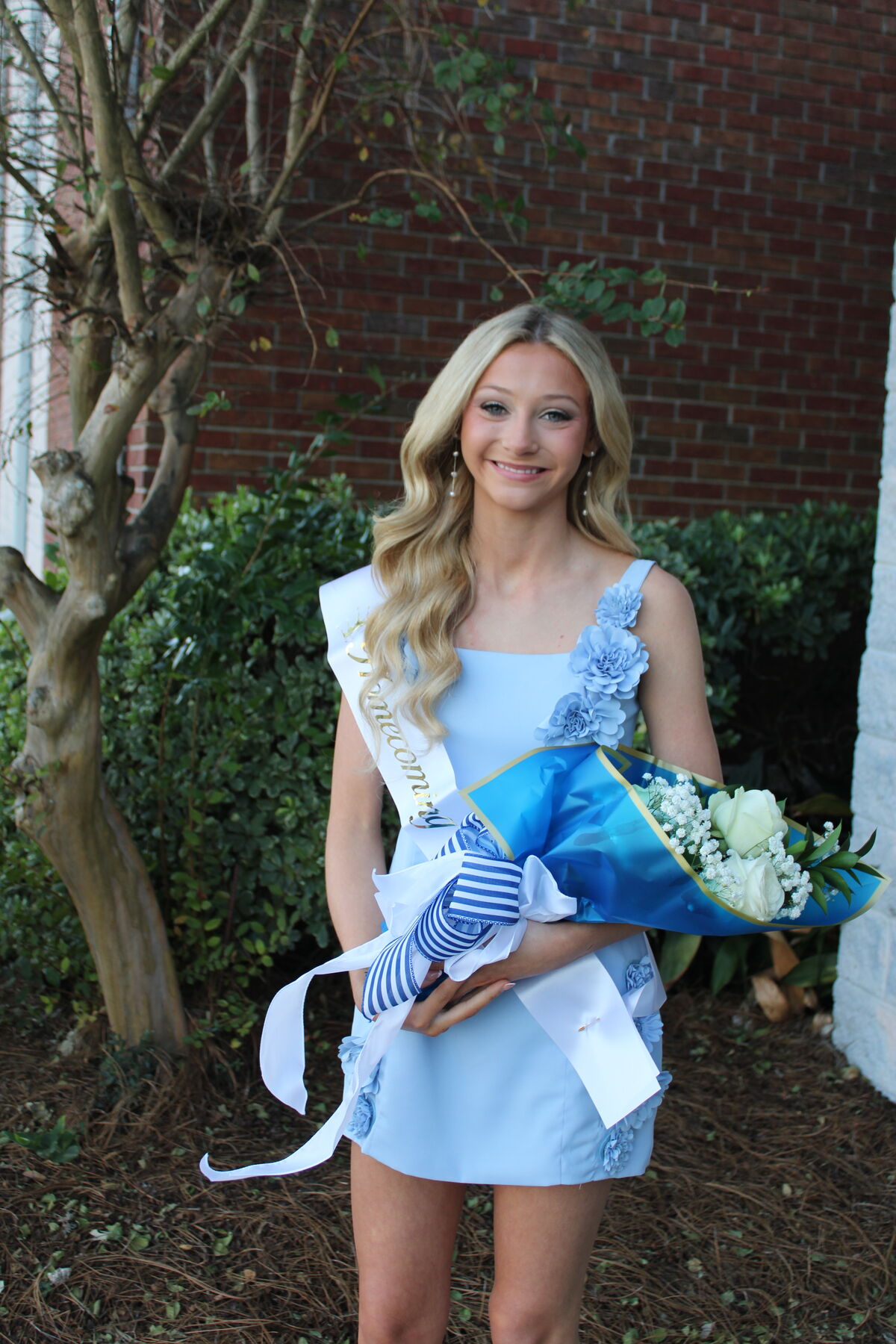 A smiling young woman in a light blue dress holds a bouquet of white roses and wears a sash reading "Homecoming".