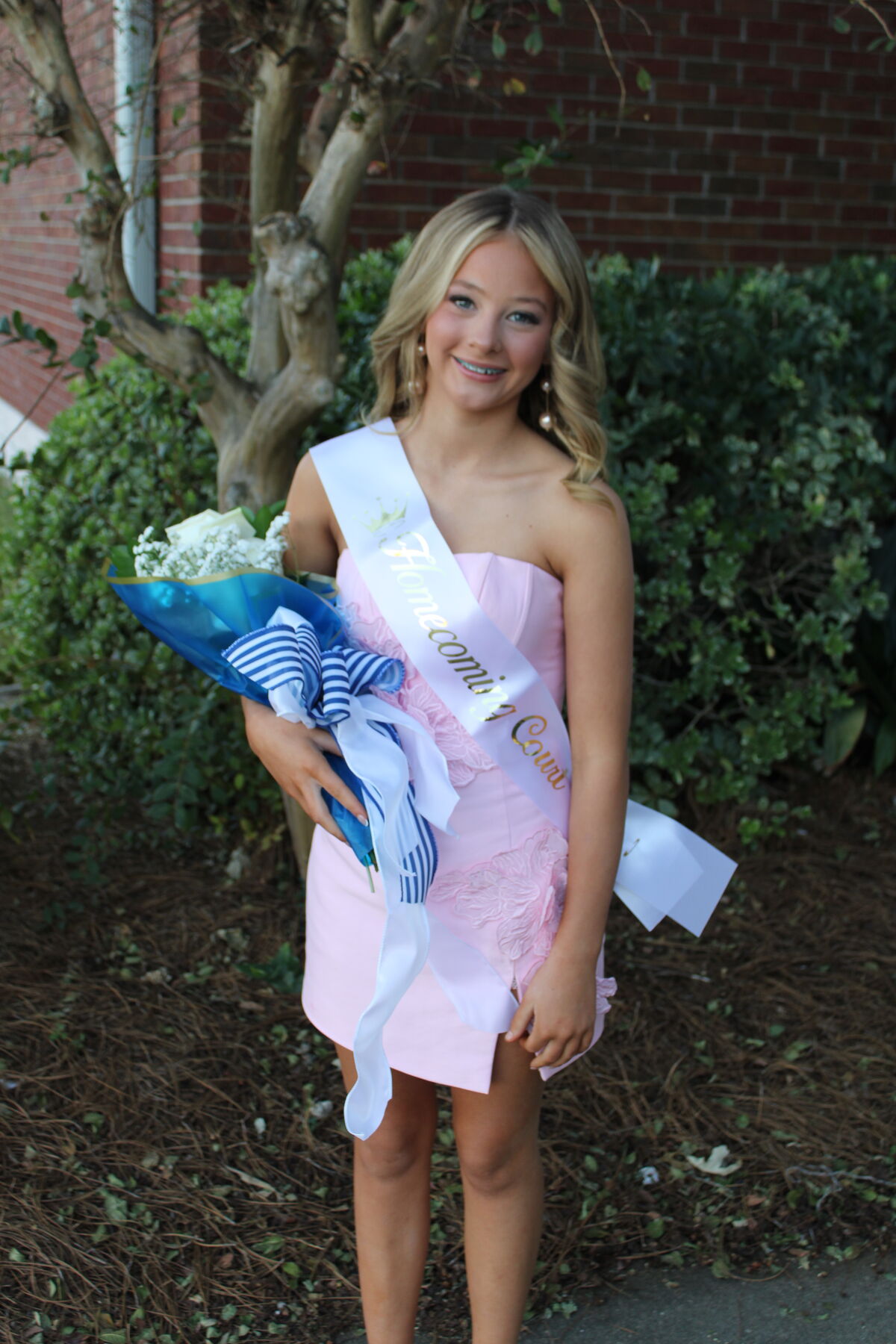 A smiling young woman in a pink dress wears a white sash with gold text reading 'Homecoming Court' while holding a bouquet of flowers wrapped in blue.