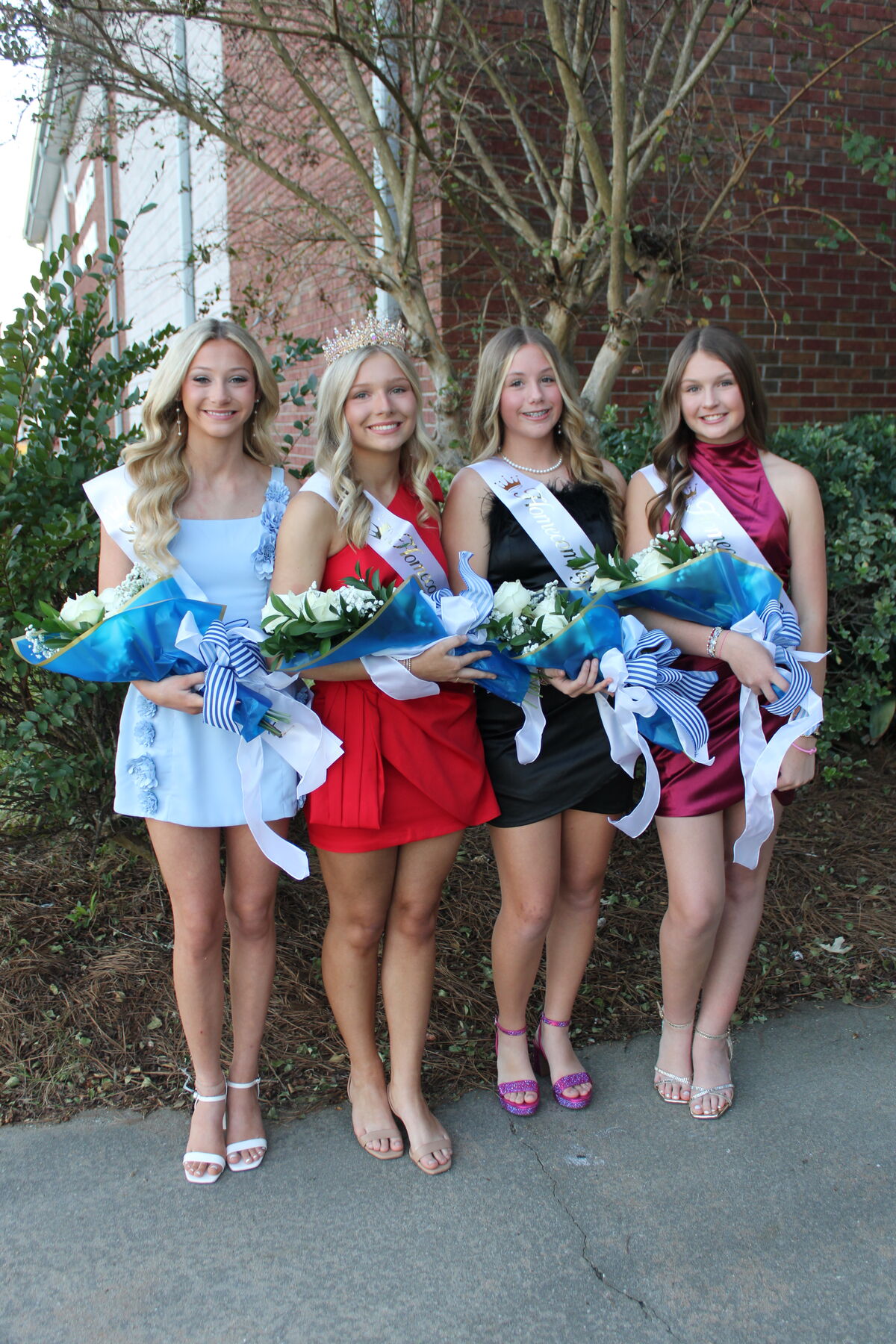 Four young women holding bouquets and wearing homecoming sashes stand outdoors near bushes and a brick building.