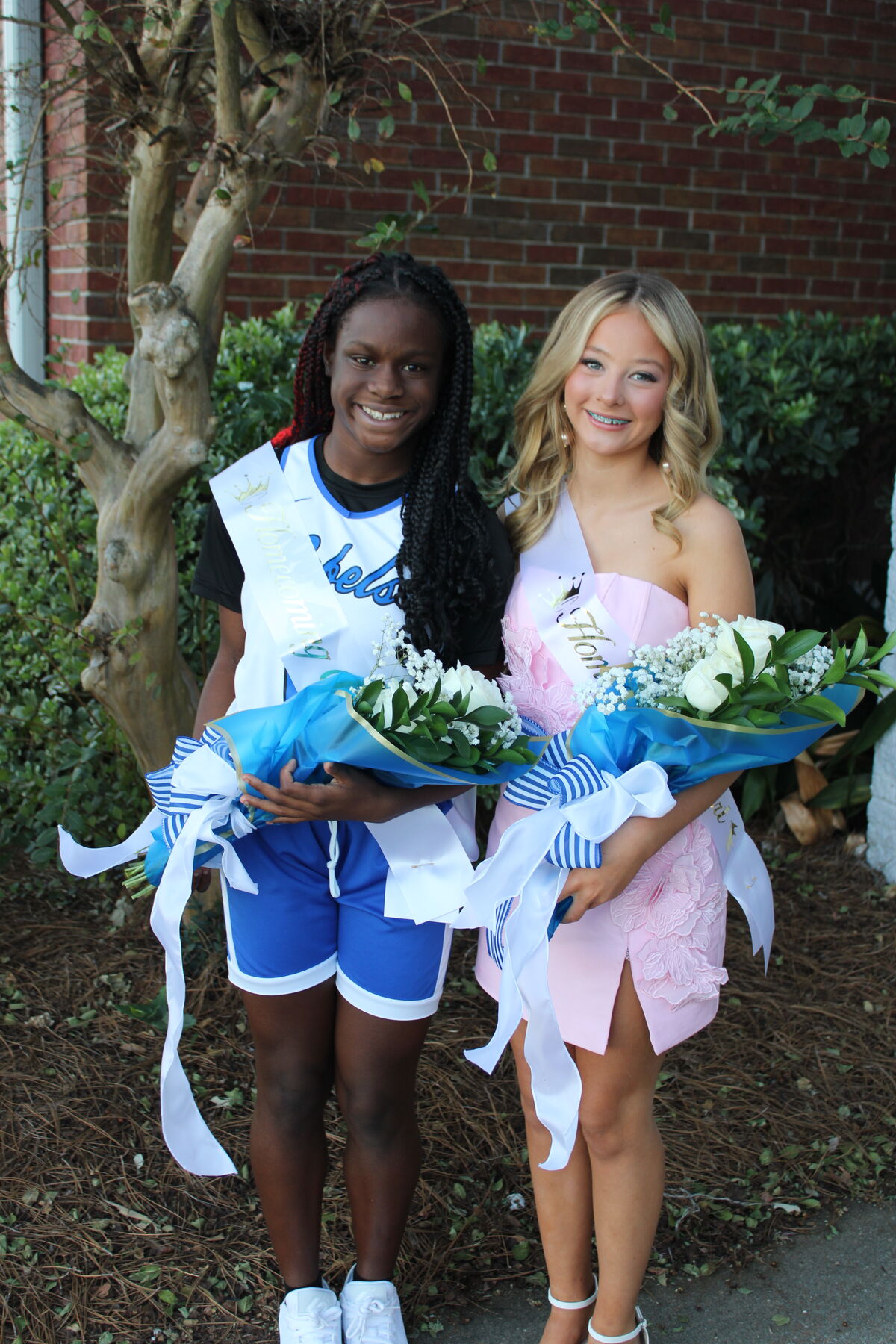 Two smiling young women wearing sashes and holding bouquets of flowers outdoors near trees and a brick wall.