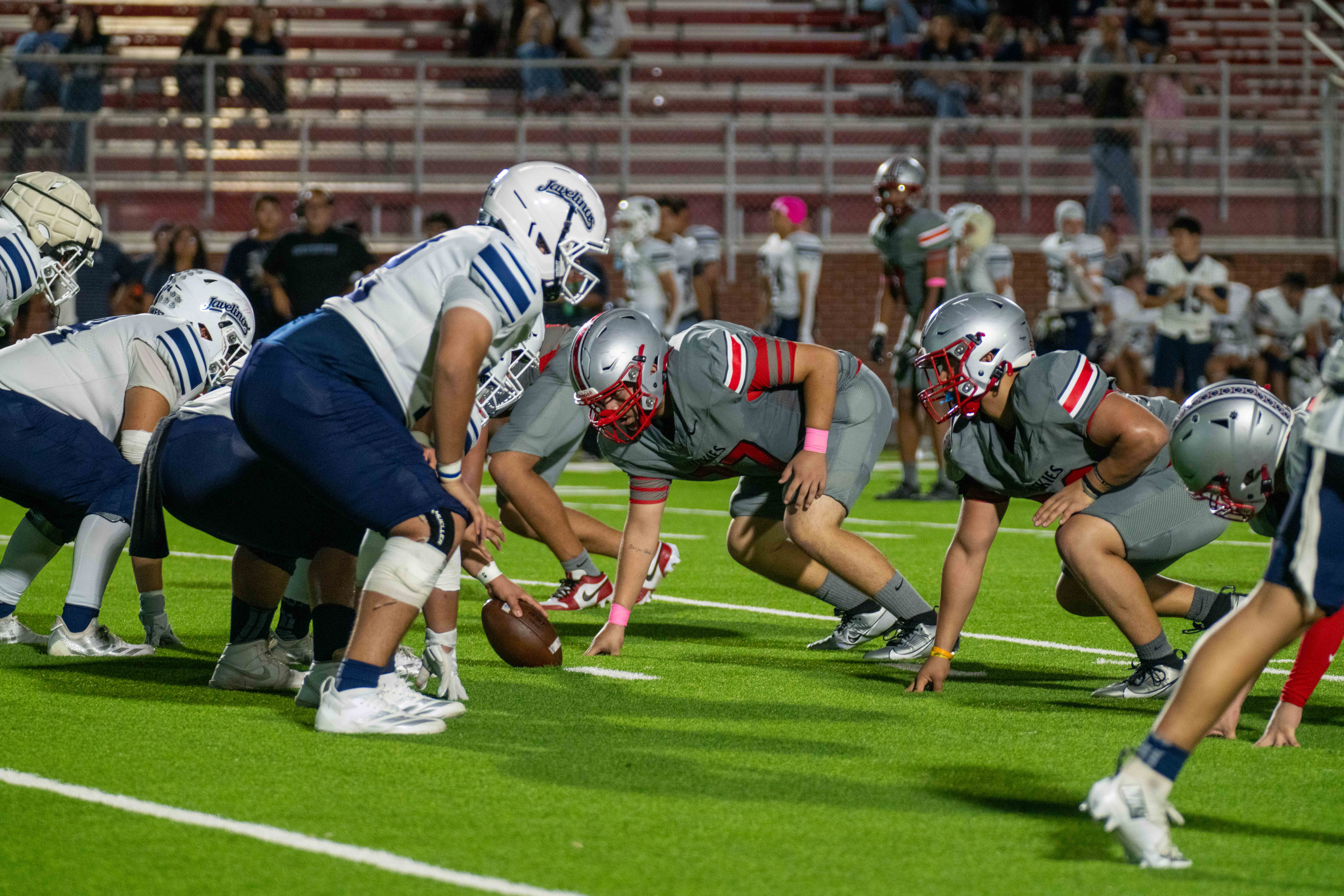 Two high school football teams line up face-to-face in a stadium at night, with the offense in white and blue uniforms and the defense in grey and red.