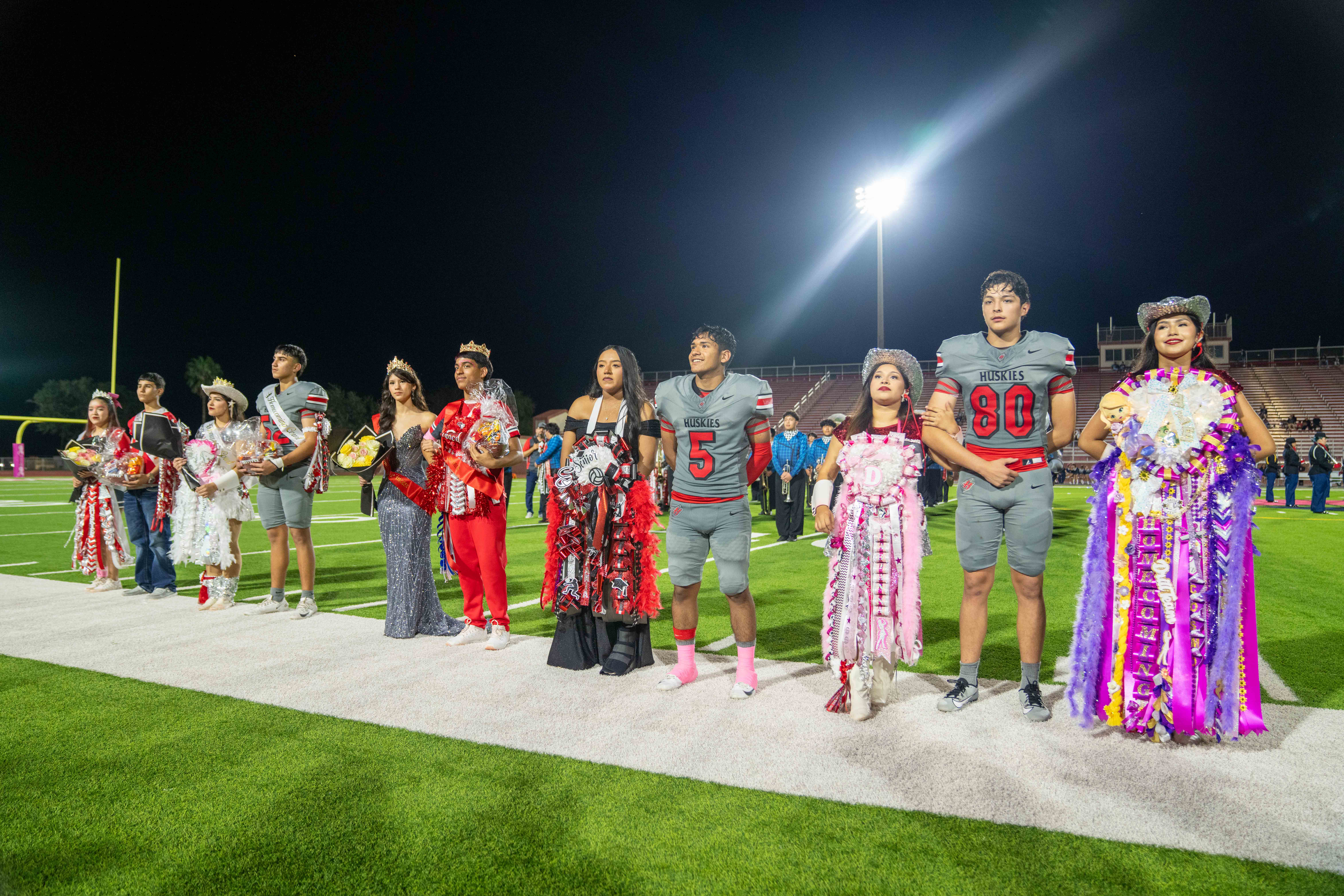 A line of high school students stands on a football field at night, participating in a homecoming court ceremony.