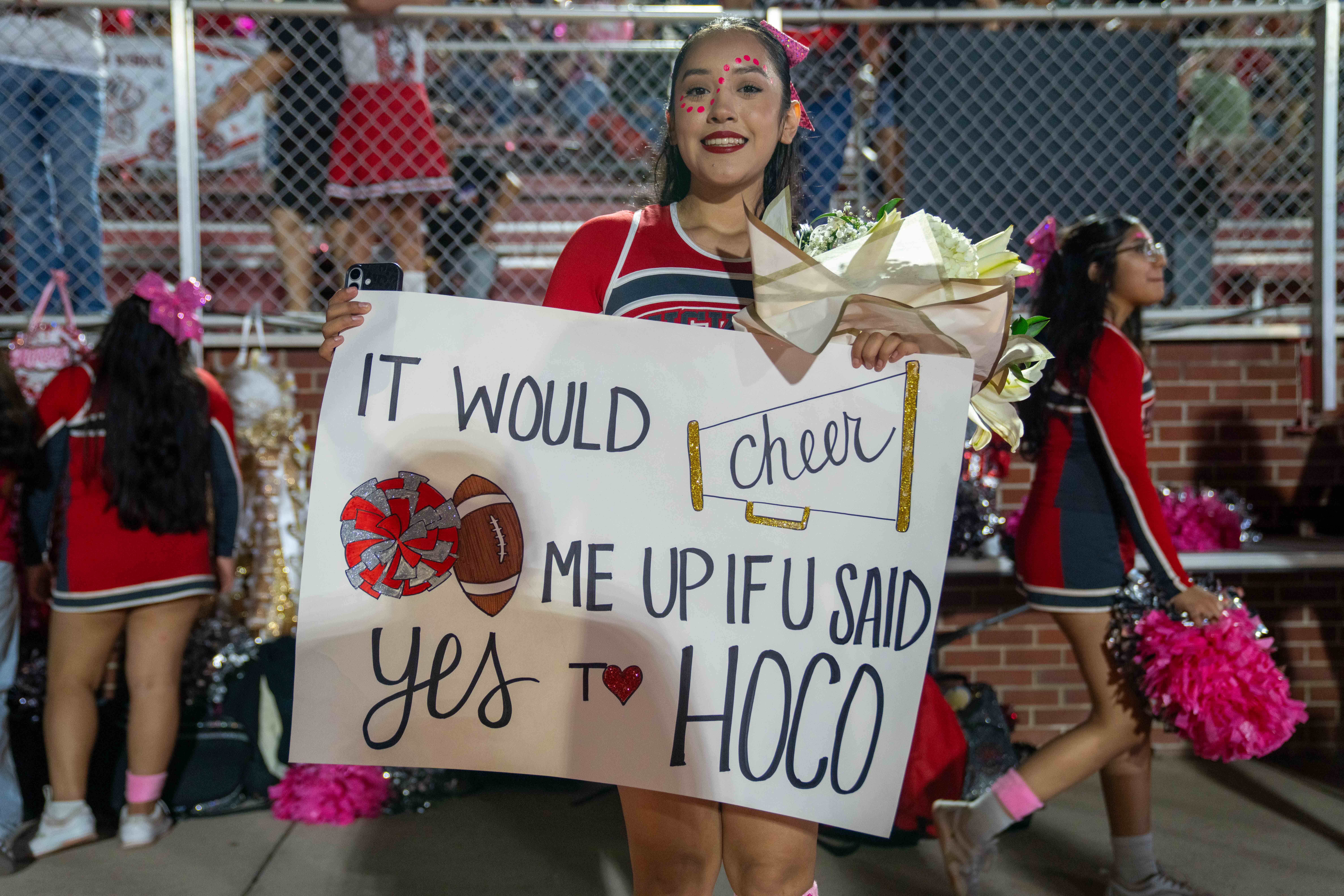 A smiling cheerleader holds a bouquet and a large sign that reads "IT WOULD CHEER ME UP IF U SAID yes TO HOCO."