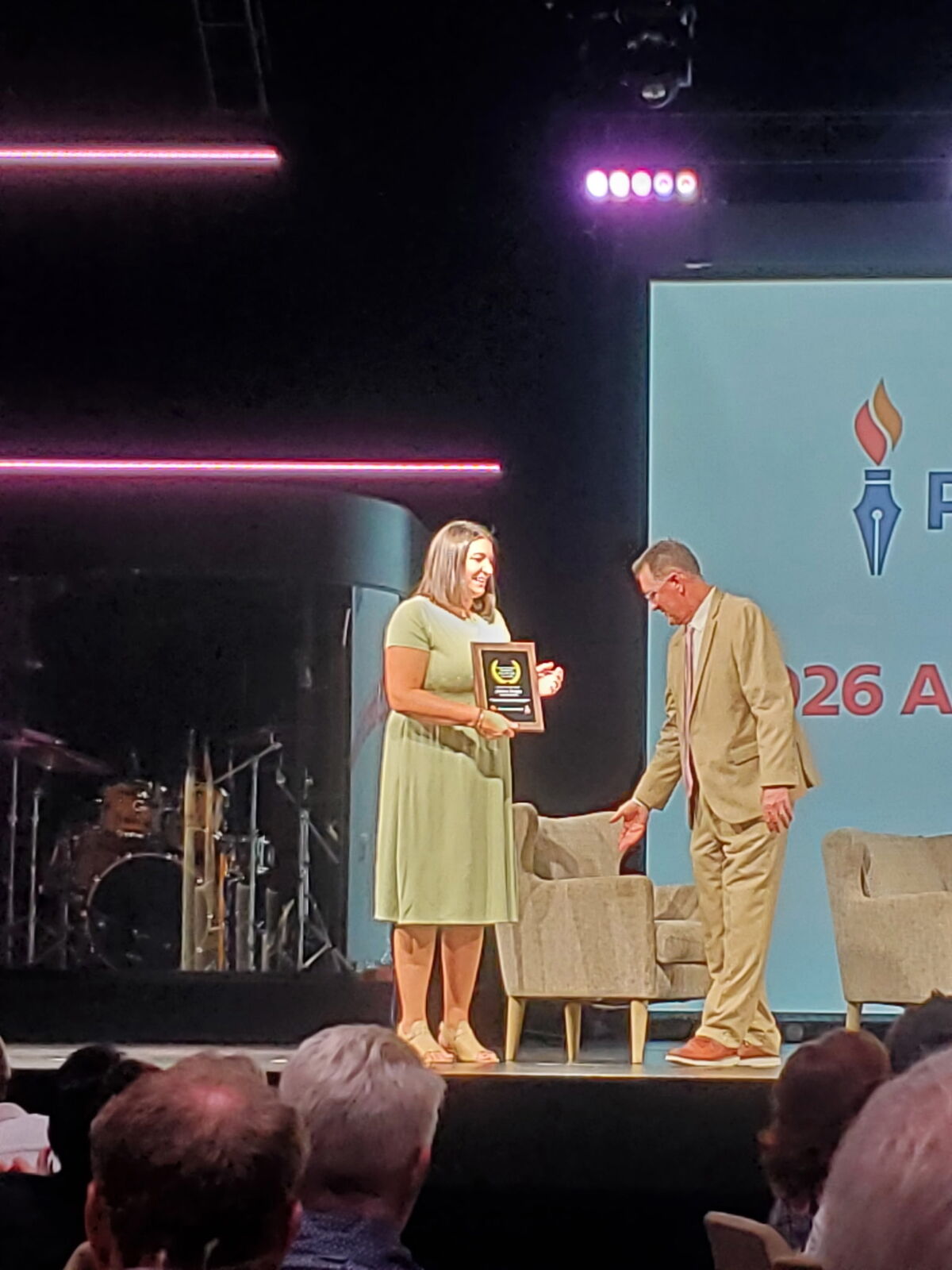 A woman in a green dress receives a framed award from a man in a tan suit on a stage.