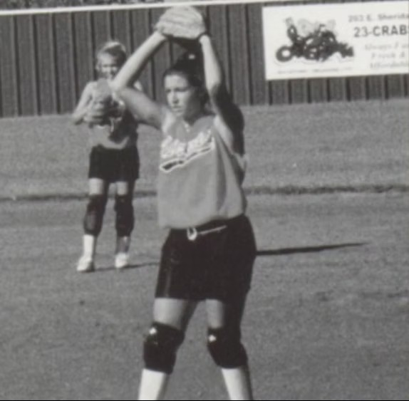A softball player winds up to pitch on a field, holding the ball above her head with another player standing in the background.