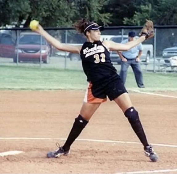 A softball pitcher in a black uniform winds up to throw a yellow ball on a dirt field.