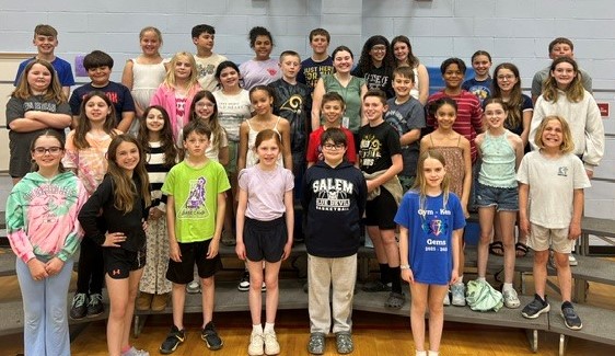 A diverse group of children and young adults pose for a photo on tiered risers in a gymnasium.