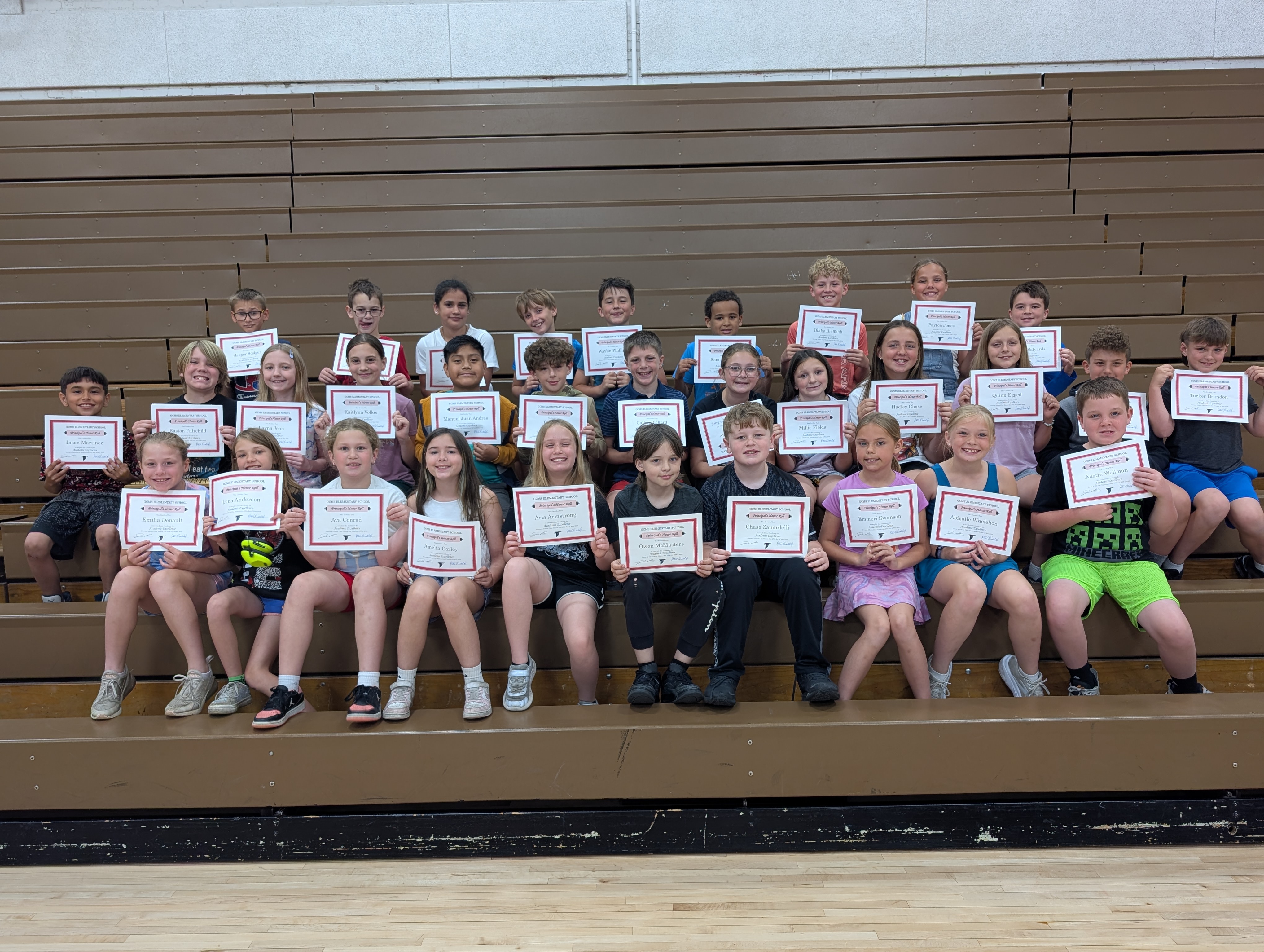 A group of students sits on bleachers, each holding a certificate from OOMS Elementary School.