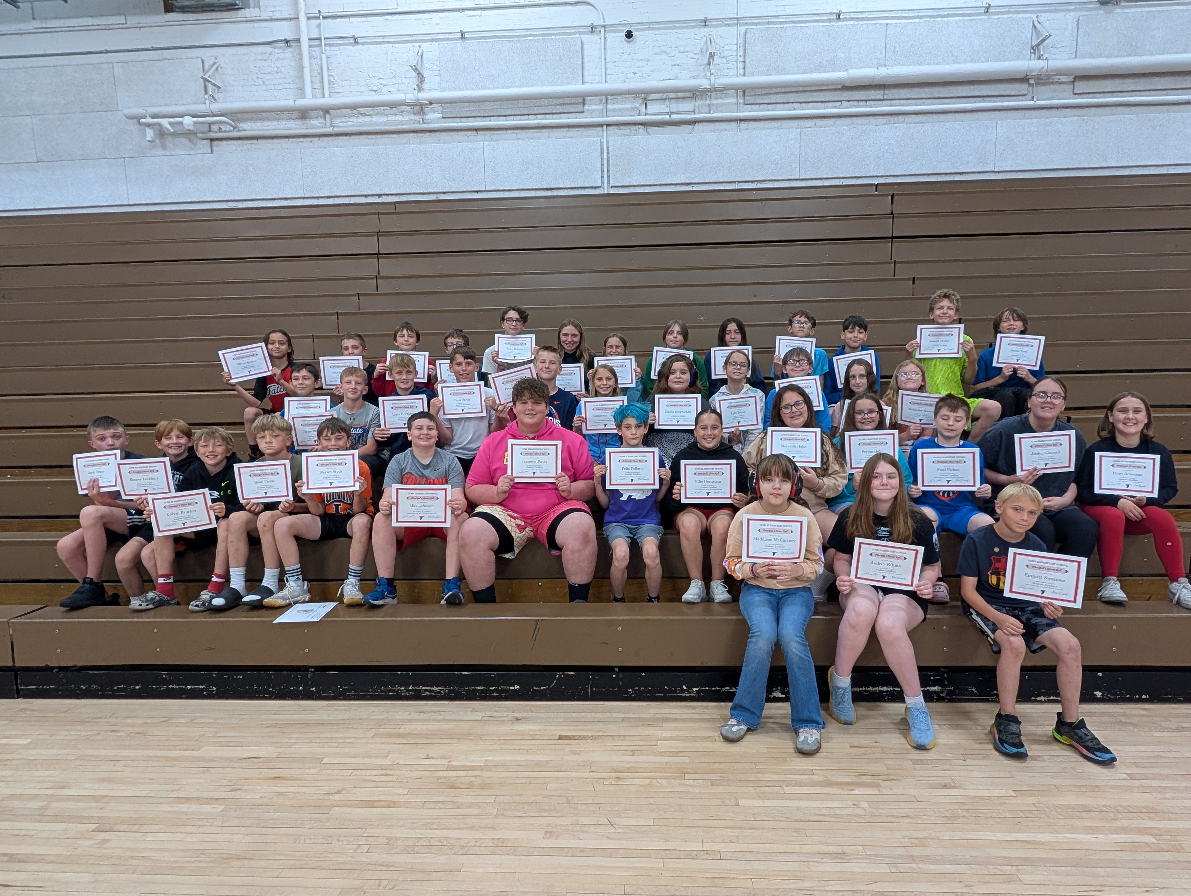 A group of students are seated on bleachers, each holding a certificate.
