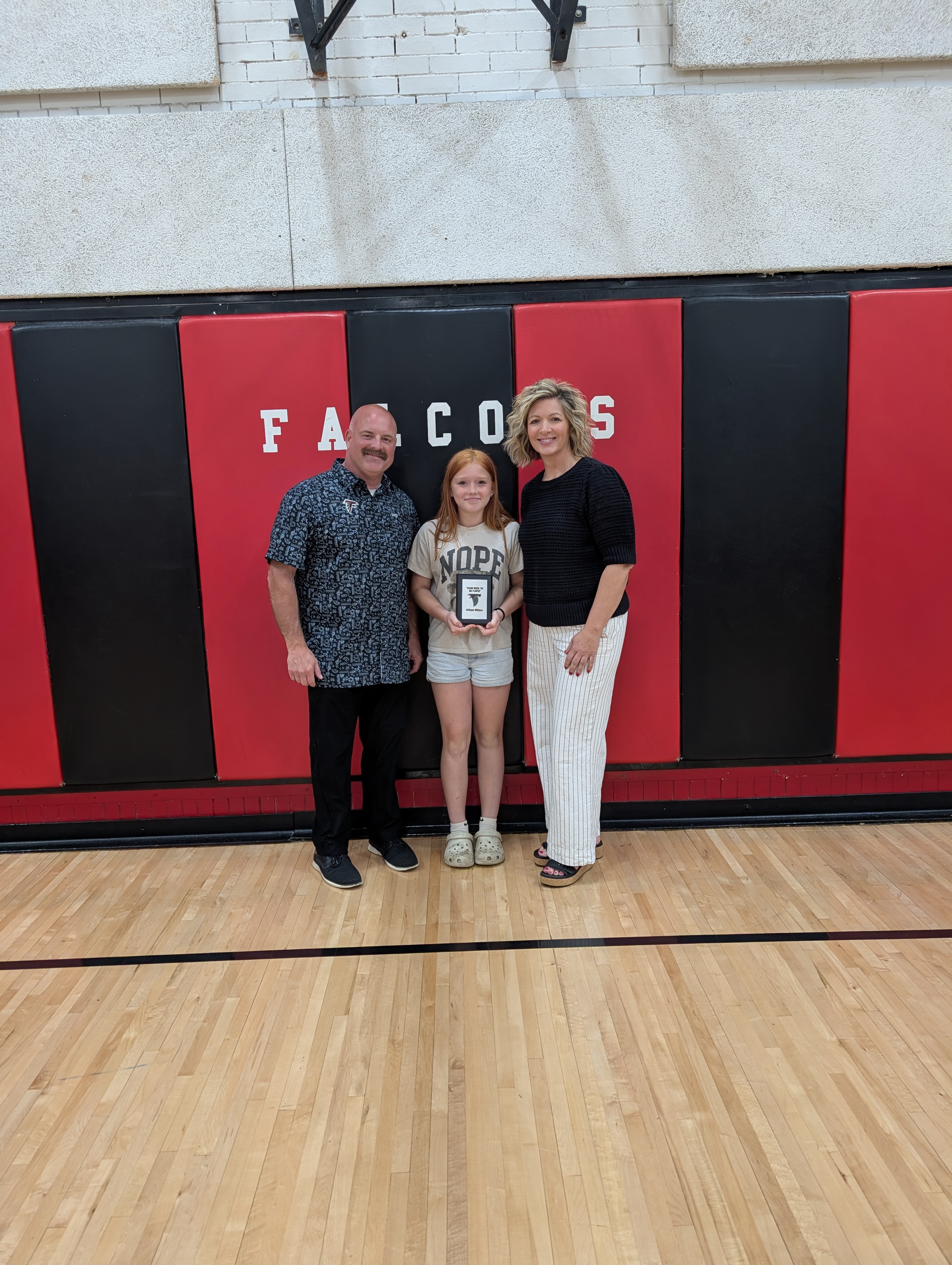 A young girl stands between two adults, holding a plaque, in front of a red and black wall with the word "FALCONS" visible.