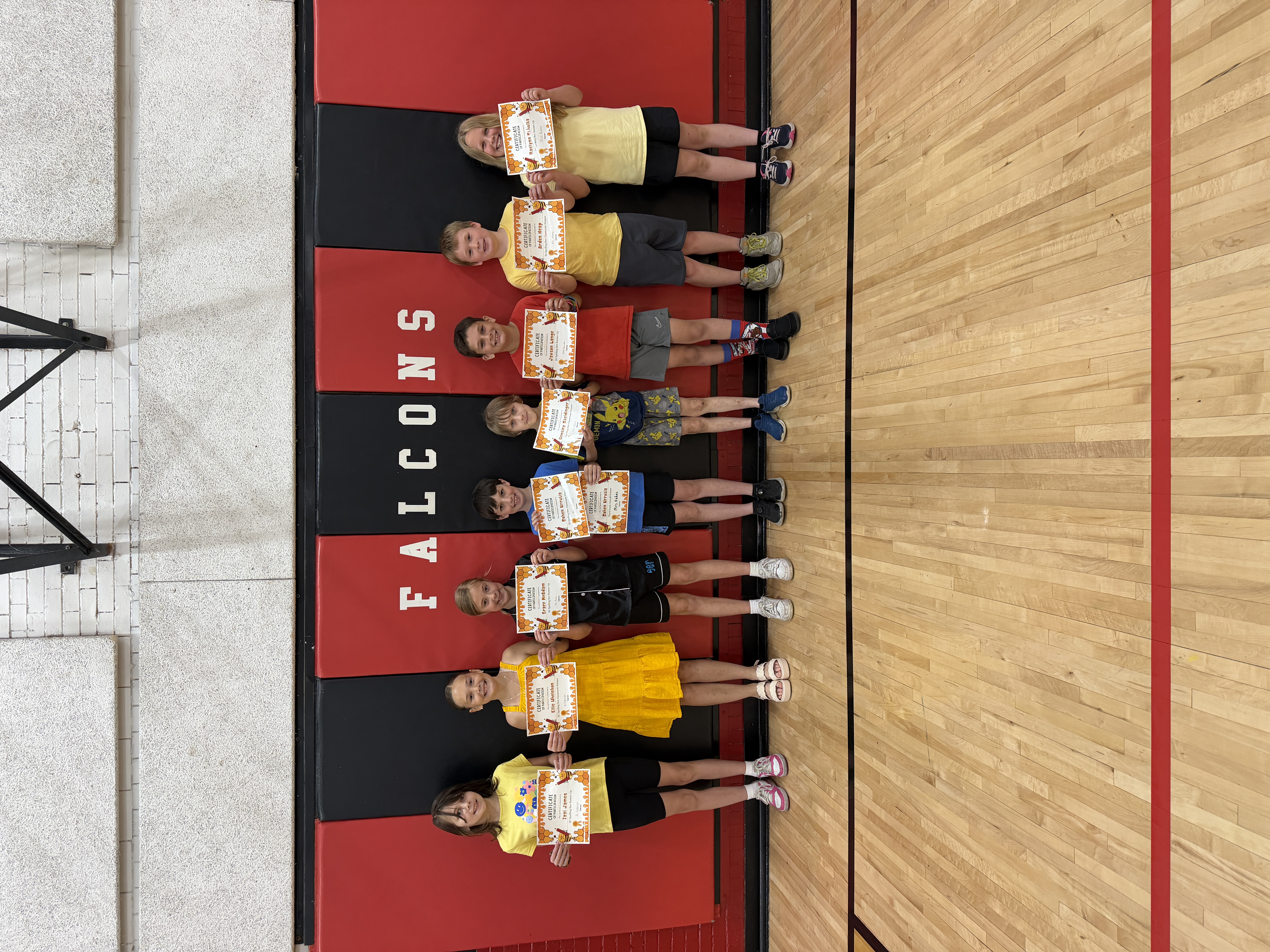 A group of children, each holding a certificate, lie on a red and black mat in a gymnasium.