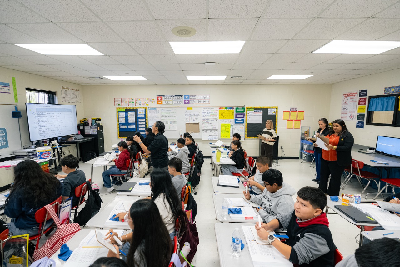 A teacher stands in front of a bright, busy classroom filled with students sitting at individual desks, reading and writing.