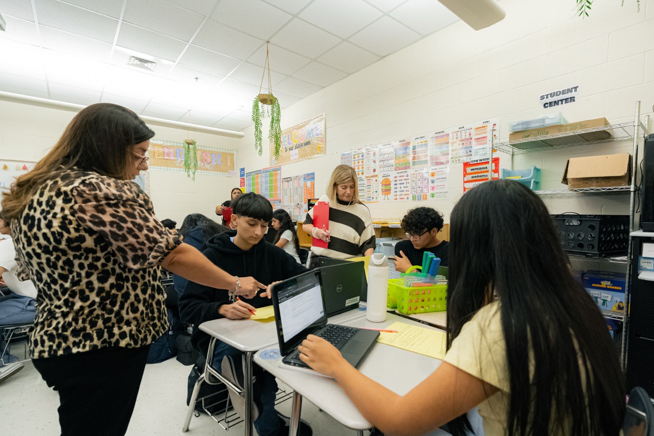 A teacher assists a student working on a laptop in a bright, colorful classroom filled with learning materials.