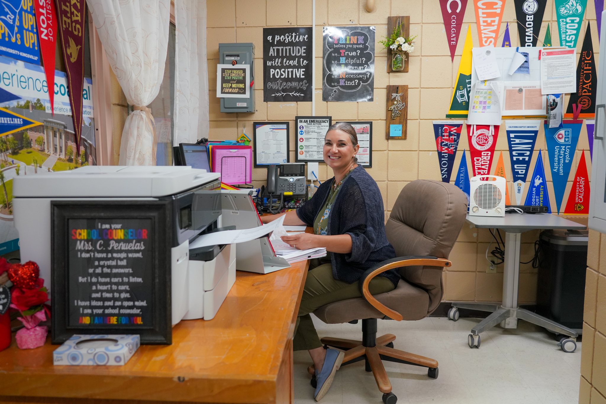 A smiling school counselor, Mrs. C. Peñuelas, sits at her desk in an office decorated with motivational posters and college pennants.