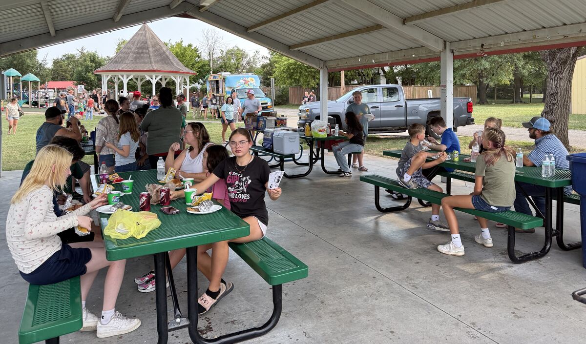 People gather under a covered pavilion, enjoying food at picnic tables with a playground and food truck in the background.
