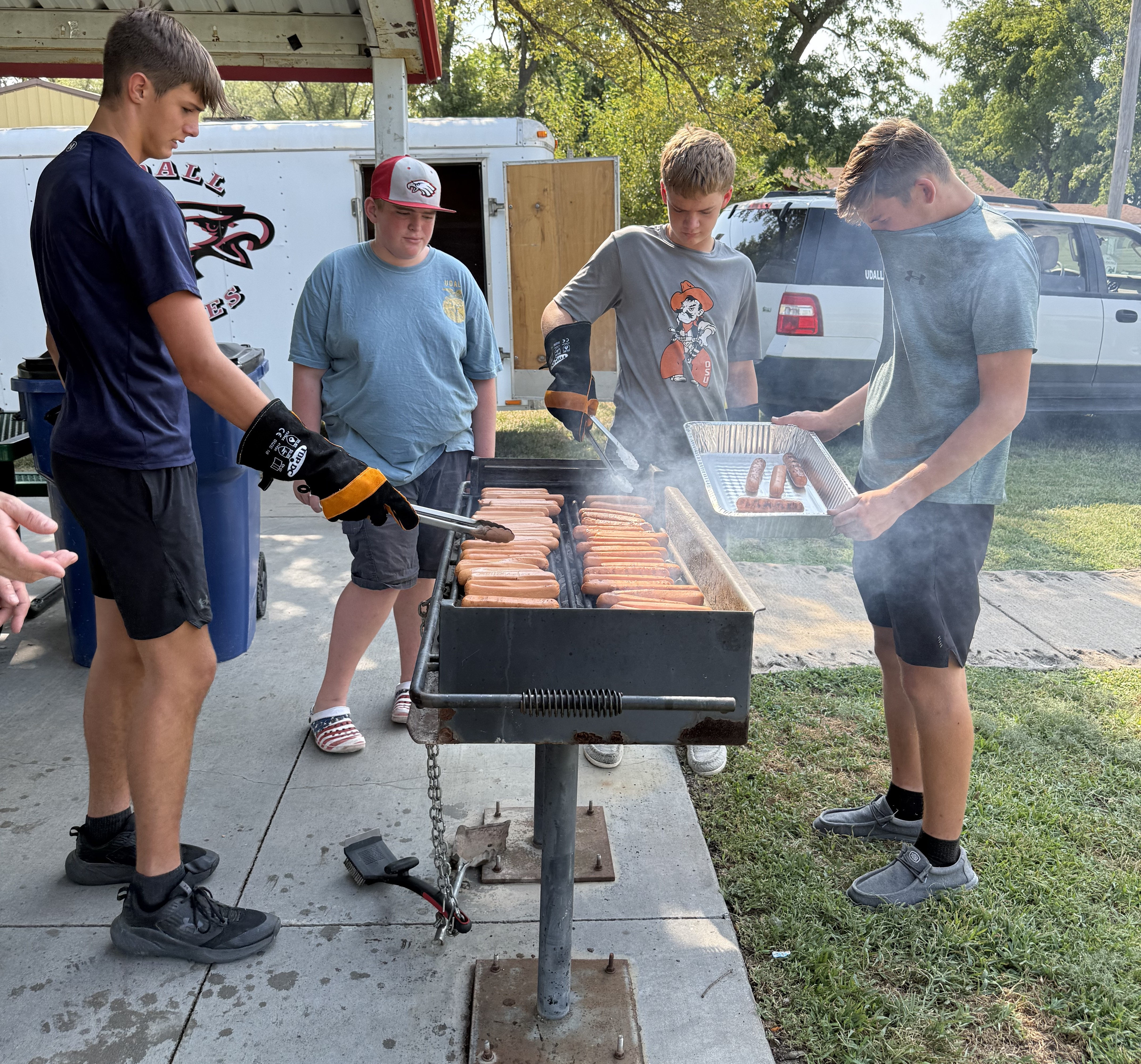 Four young people are grilling hot dogs outdoors on a sunny day.