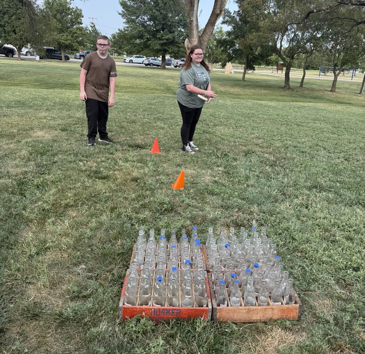 Two young people stand in a grassy park, with a game setup of glass bottles in crates.