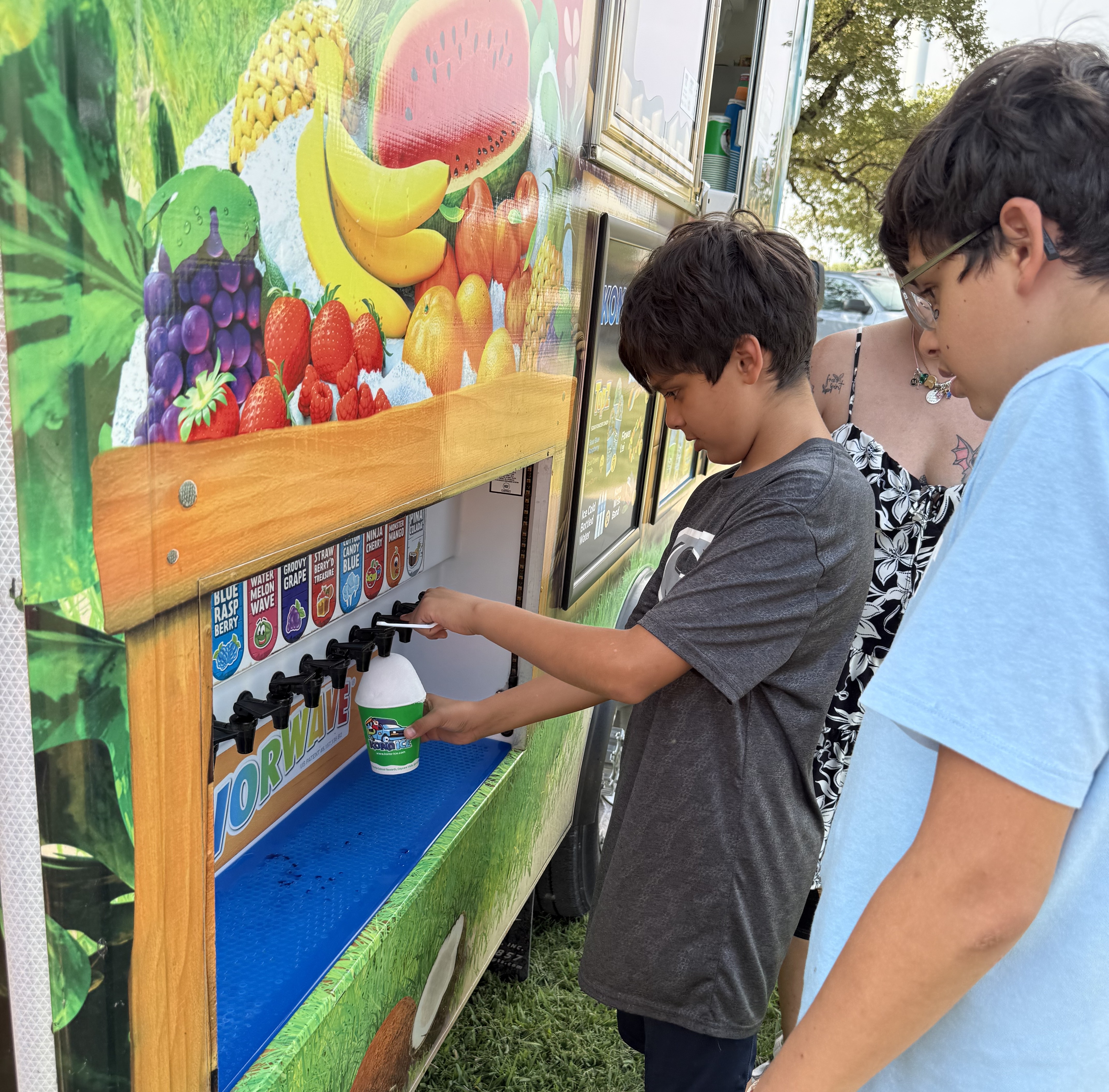 A boy uses a dispenser on a colorful Kona Ice truck to fill a cup with shaved ice.