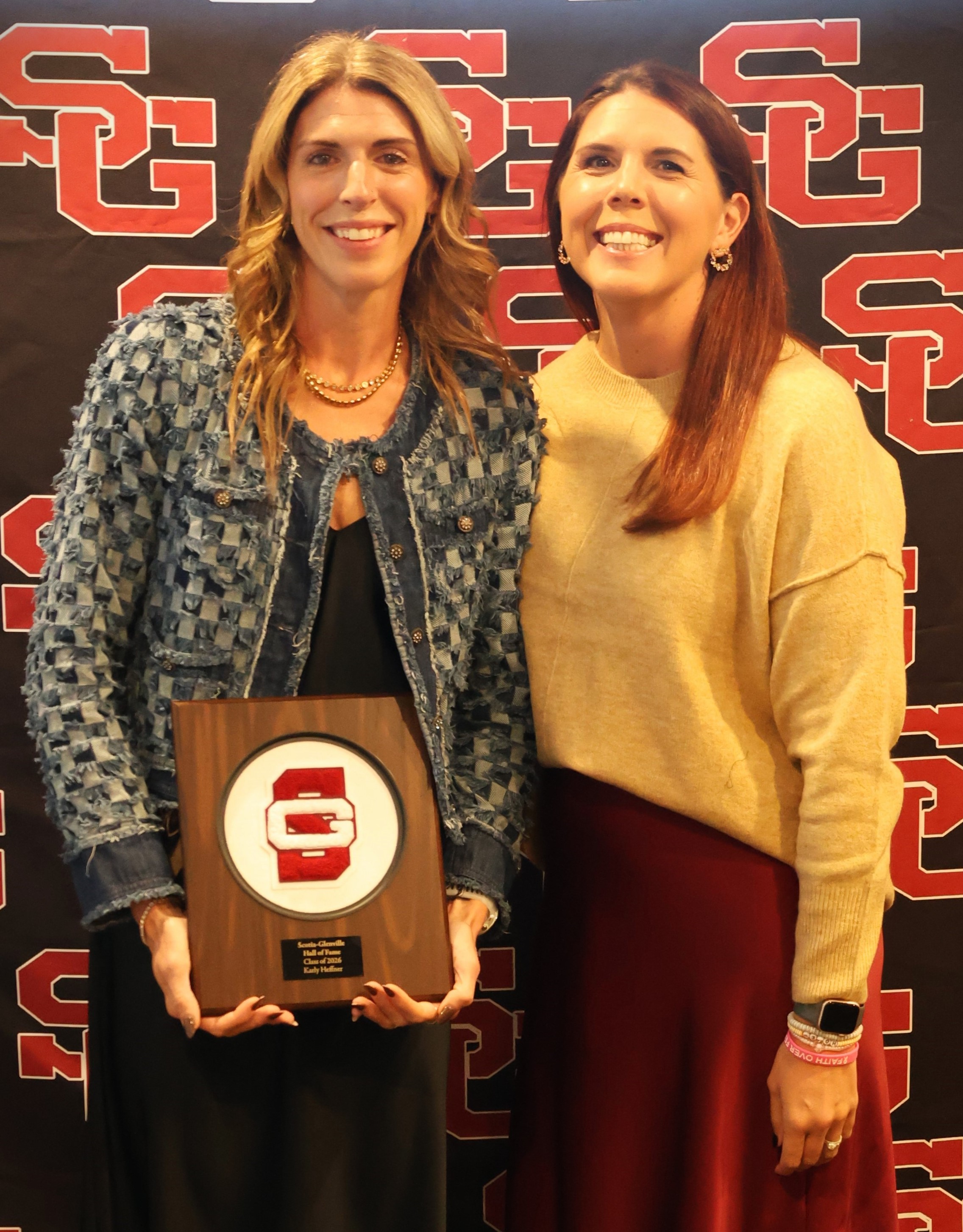 Two smiling women stand in front of a red and black patterned backdrop, with one holding a commemorative wooden plaque.
