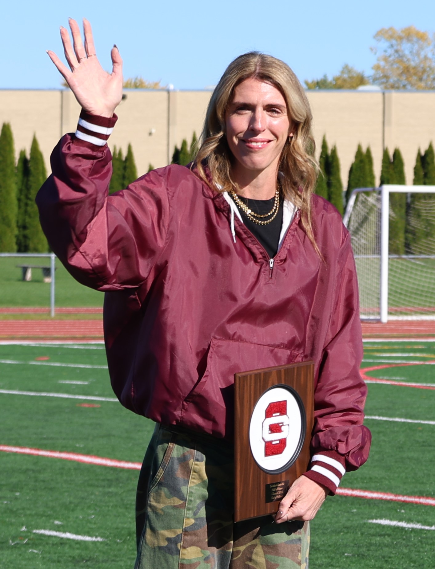 A smiling woman wearing a maroon jacket waves and holds a commemorative plaque on an outdoor sports field.