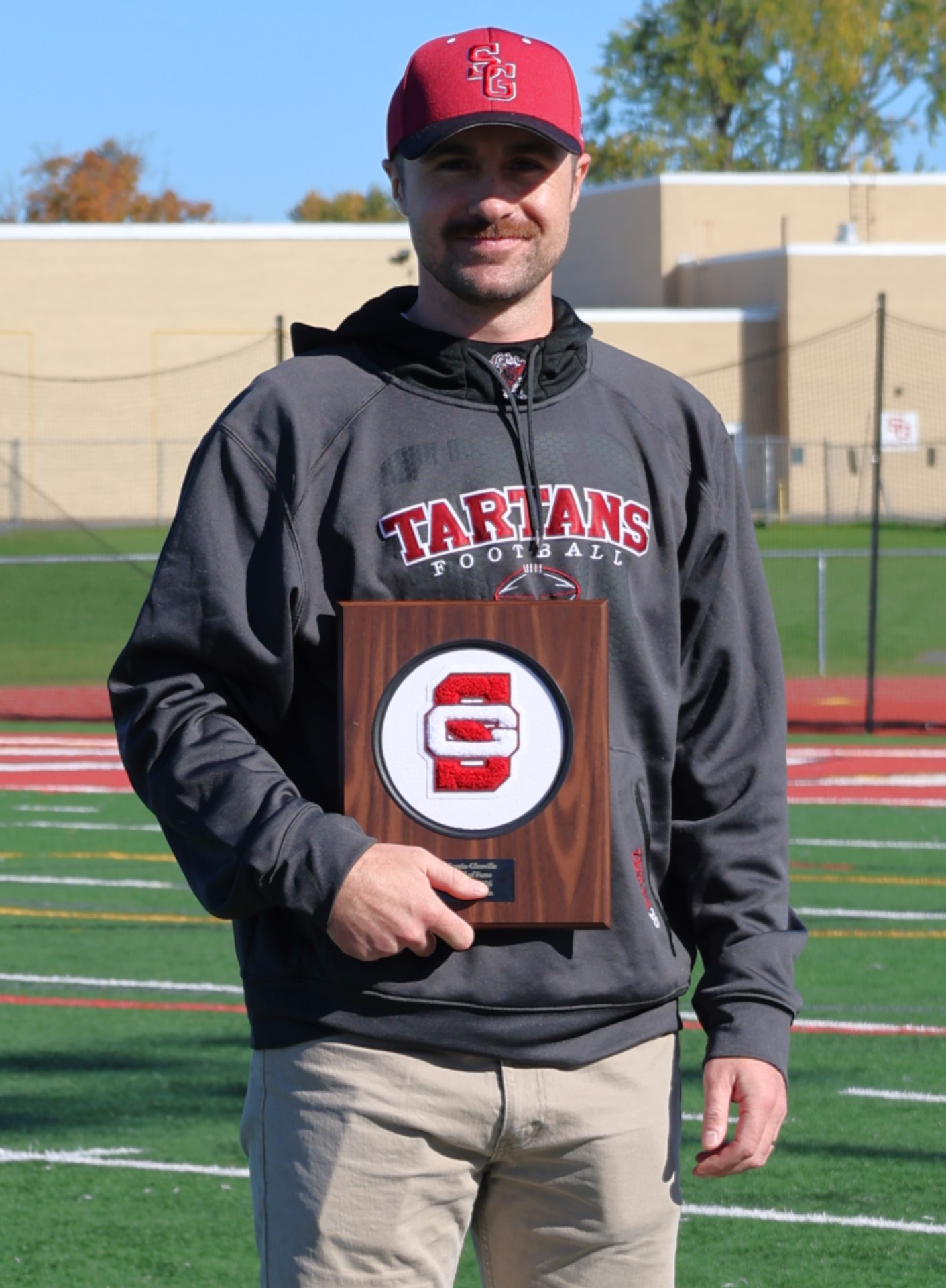 A man wearing a red baseball cap and a dark Tartans Football hoodie holds a wooden award plaque outdoors on a sports field.