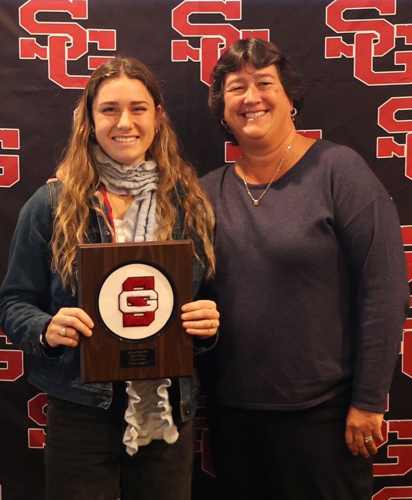 Two smiling women stand in front of a red and black patterned backdrop, holding a wooden plaque.
