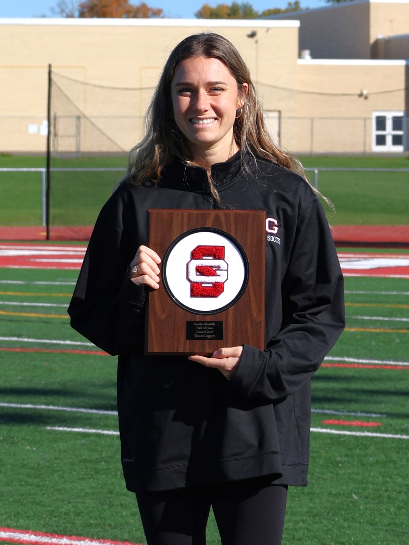 A smiling woman holds a wooden award plaque with a red and white "SG" logo on a sports field.