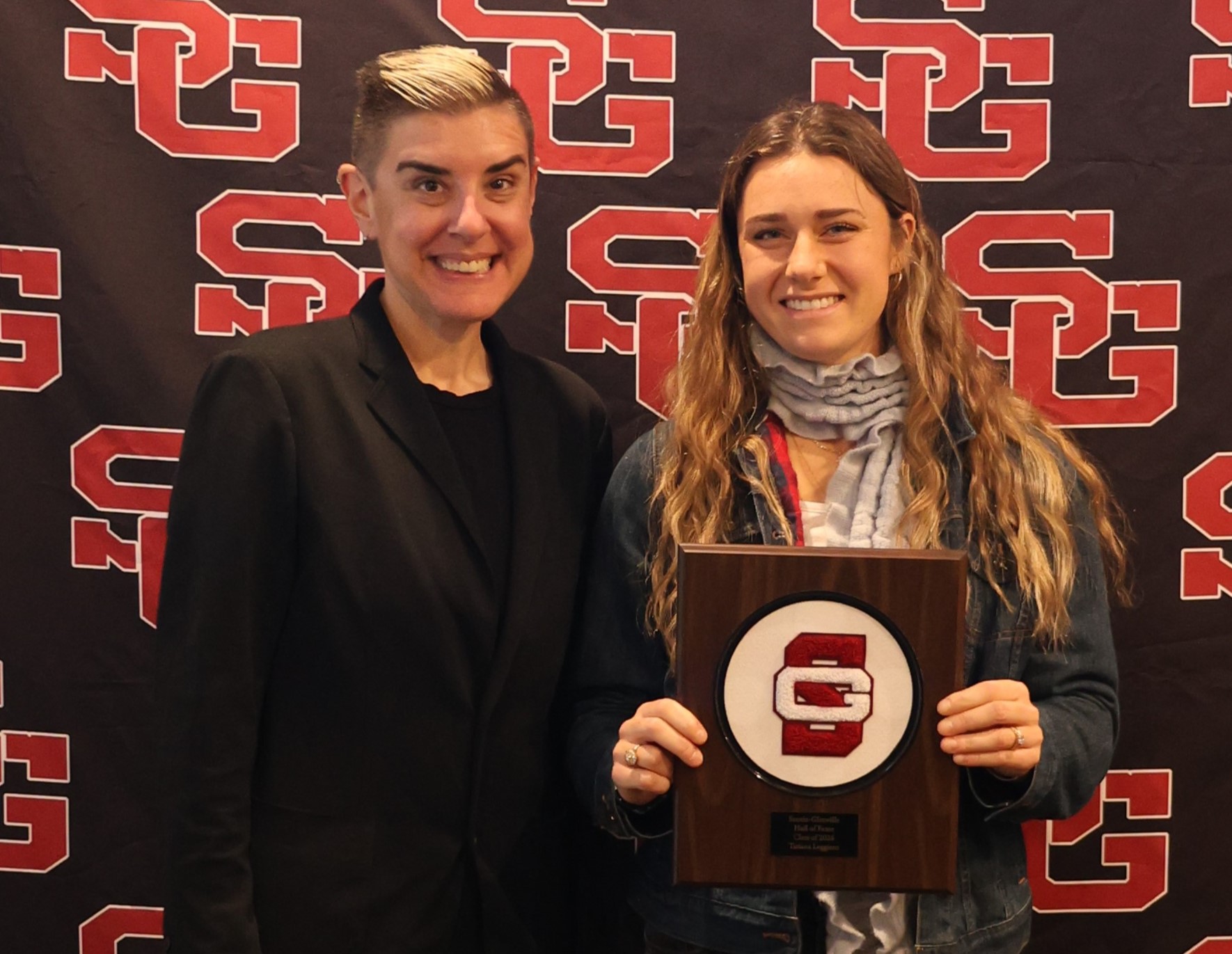 Two smiling people stand in front of a red and black patterned backdrop, holding a wooden plaque featuring a red "SC" logo.
