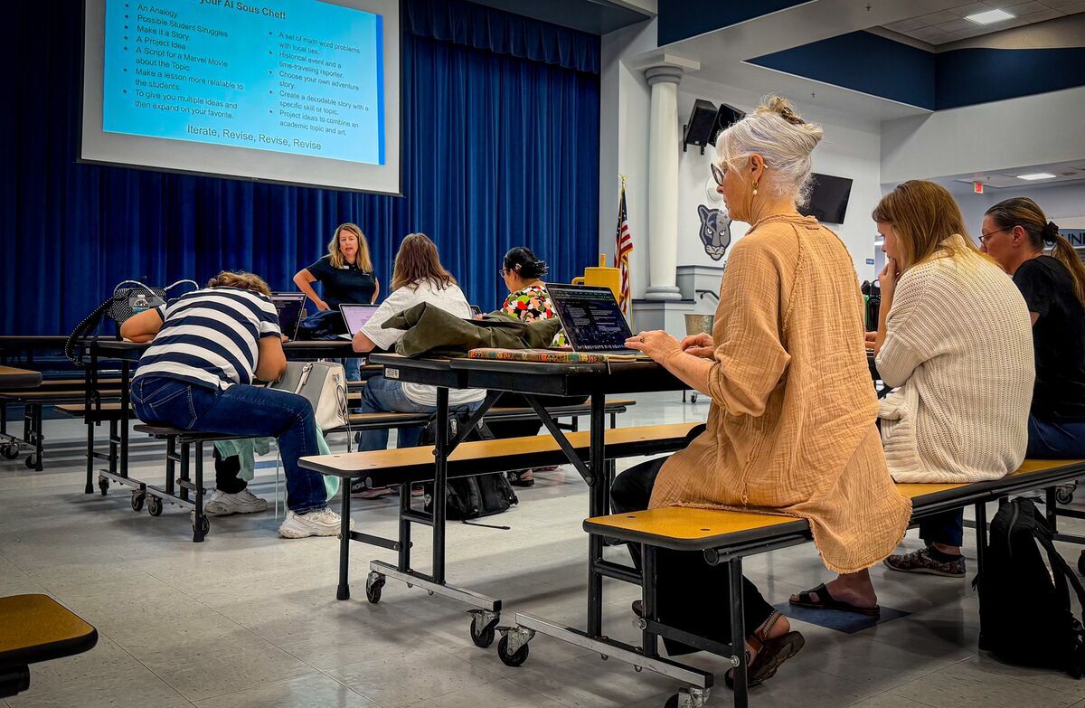 A teacher presents ideas on a projector screen to students working on laptops at cafeteria tables.