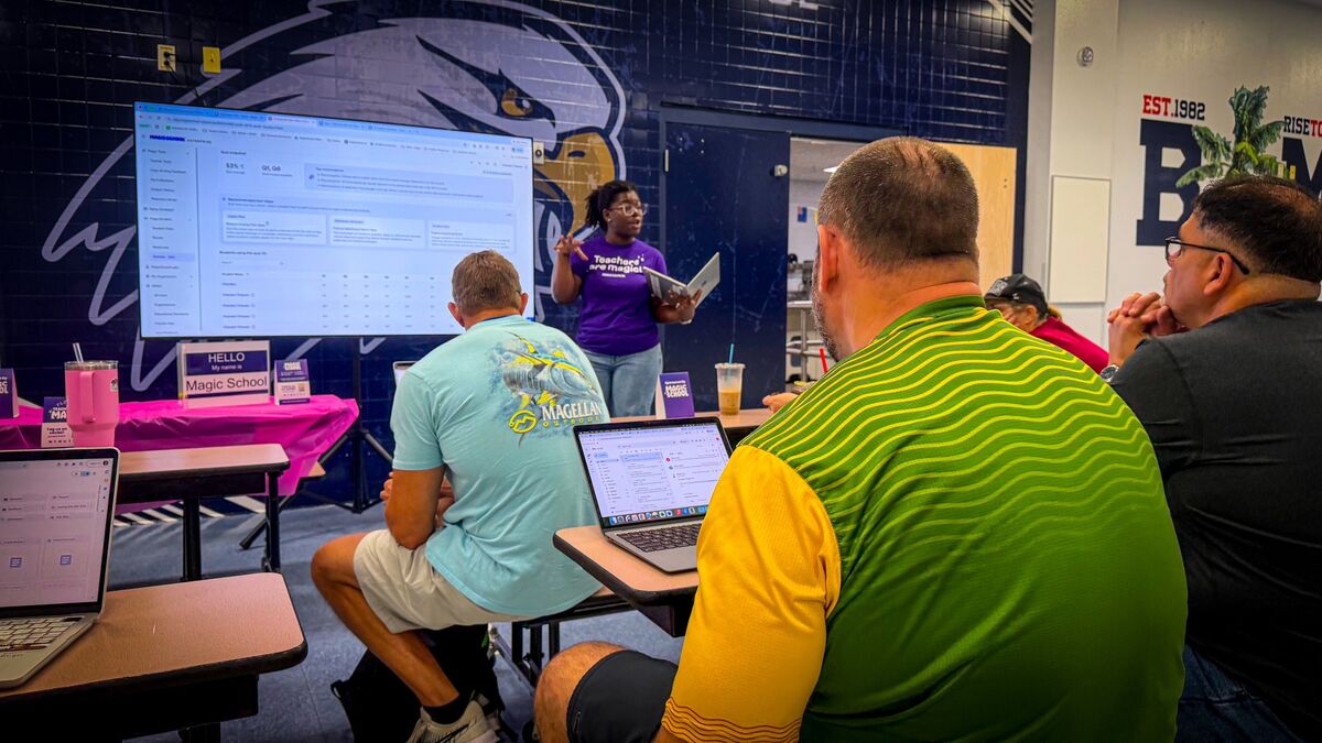 A presenter in a purple shirt speaks to an audience seated at desks with laptops, in front of a large screen displaying data.