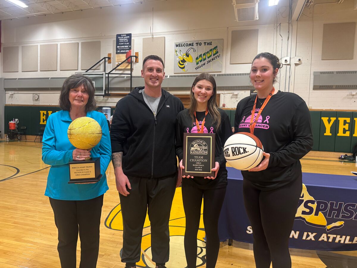 Four people stand in a gymnasium, holding a basketball trophy, a plaque, and a basketball.