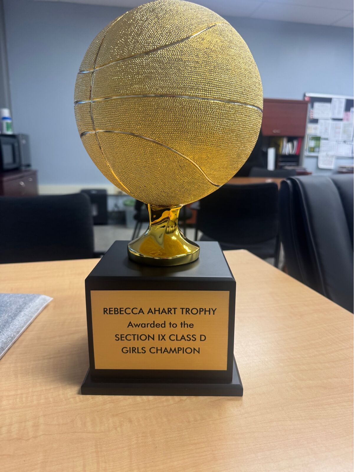 A golden basketball trophy sits on a wooden desk, engraved with 'REBECCA AHART TROPHY Awarded to the SECTION IX CLASS D GIRLS CHAMPION'.