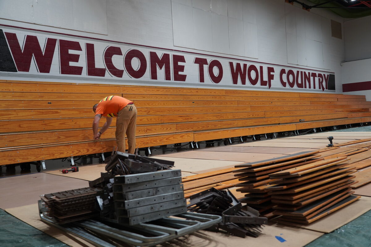 A worker in an orange shirt and tan pants adjusts bleachers in a gymnasium with "WELCOME TO WOLF COUNTRY" painted on the wall.