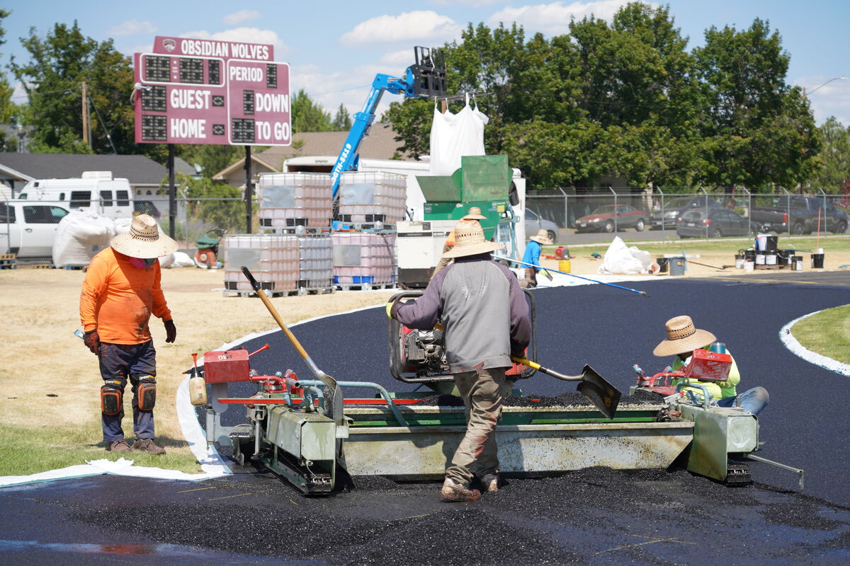 Workers in straw hats operate asphalt paving equipment on a sunny day, with a scoreboard in the background reading 'OBSIDIAN WOLVES'.