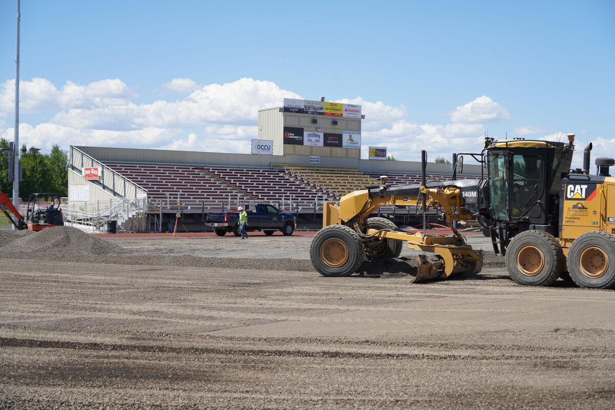 A yellow Caterpillar grader operates on a gravel surface in front of stadium bleachers.