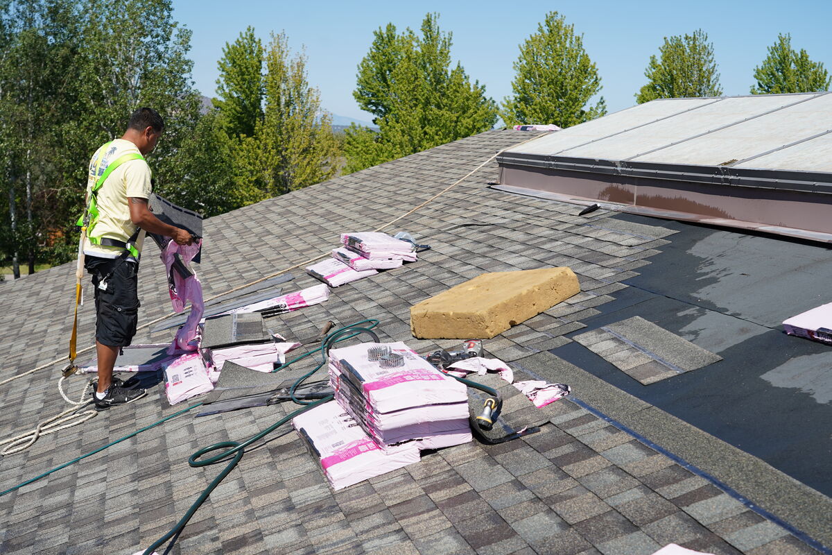 A roofer in a safety harness works on a shingled roof, surrounded by stacks of pink-wrapped roofing materials.