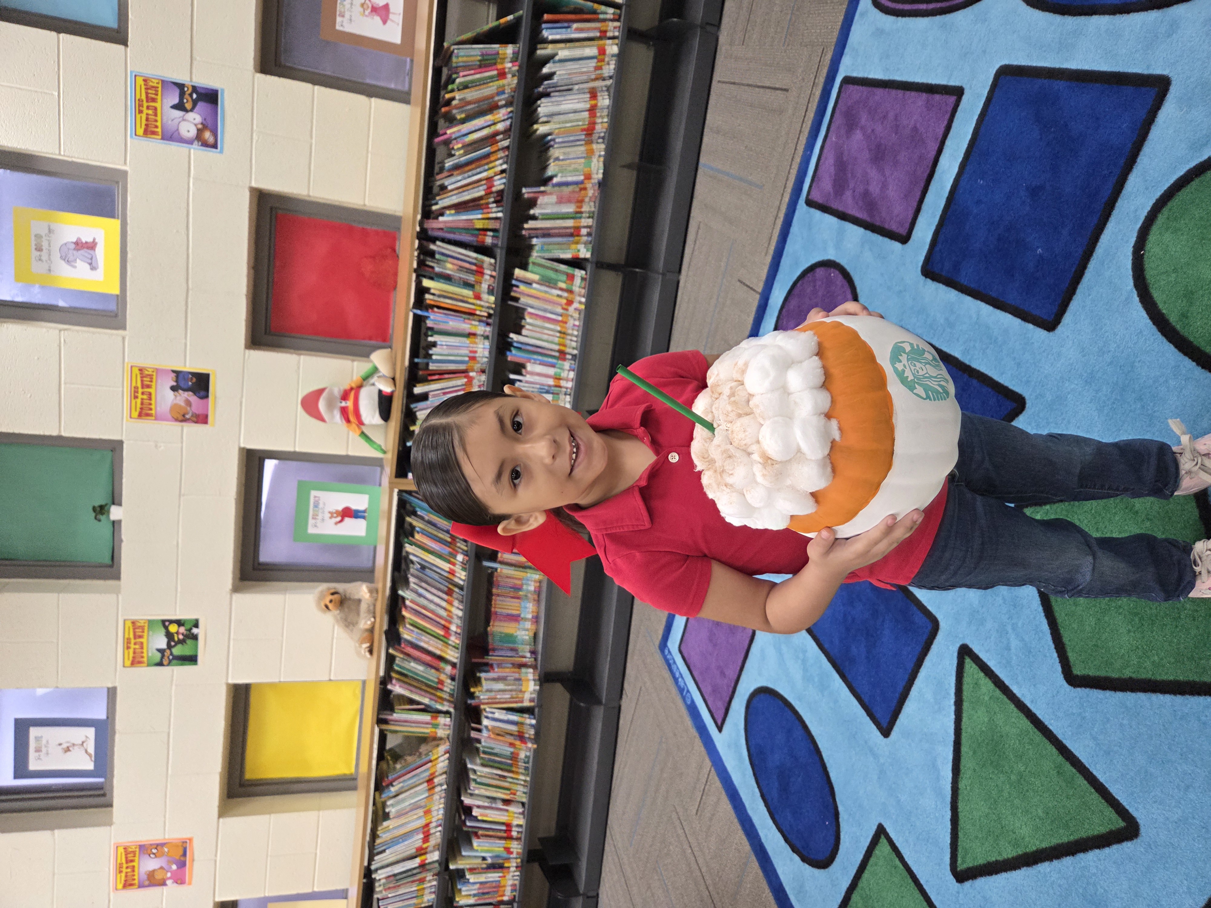 A child proudly holds a pumpkin decorated to look like a Starbucks Frappuccino in a school library.