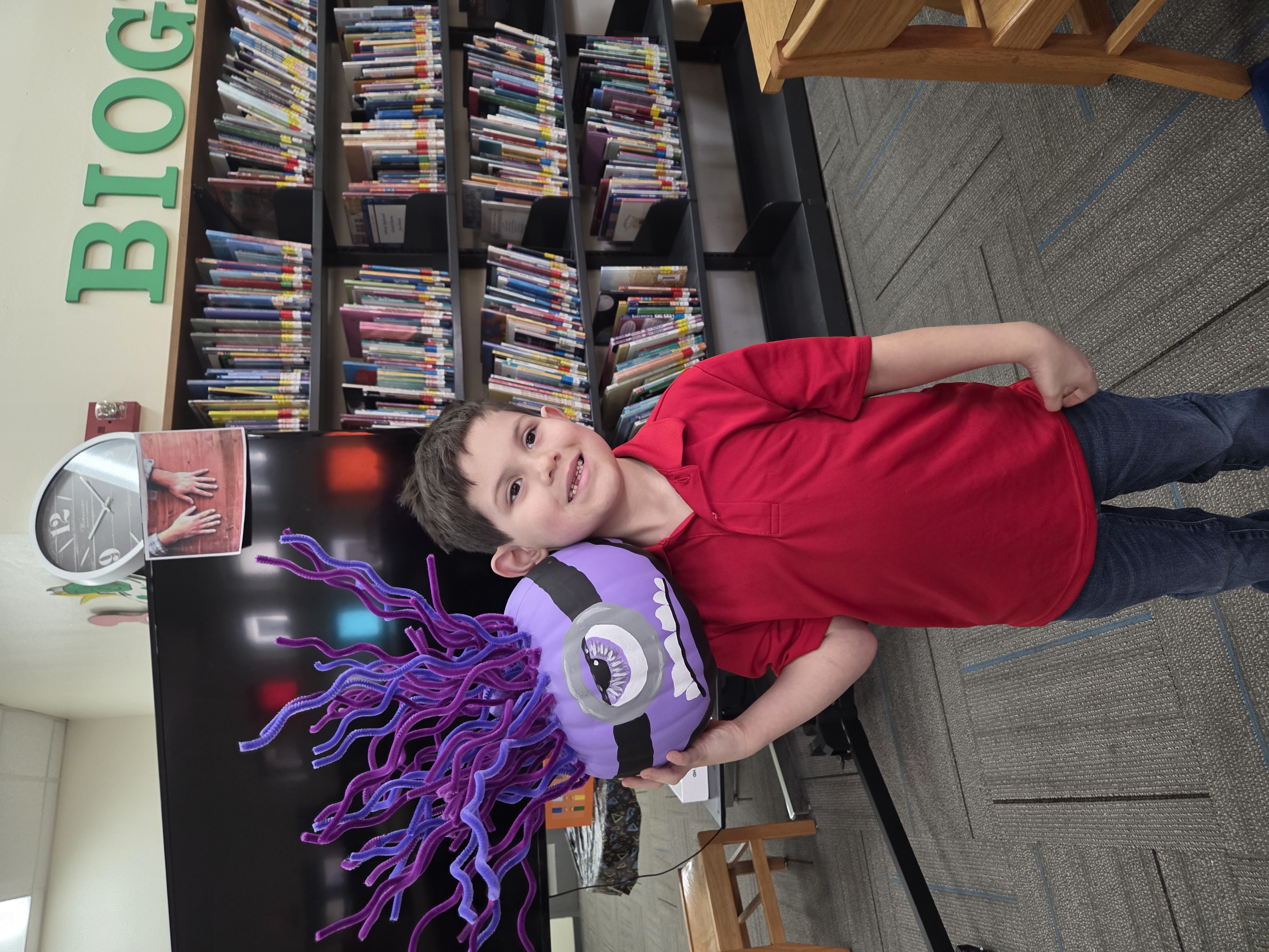 A smiling child holds a creative purple monster craft made from a pumpkin in a library.