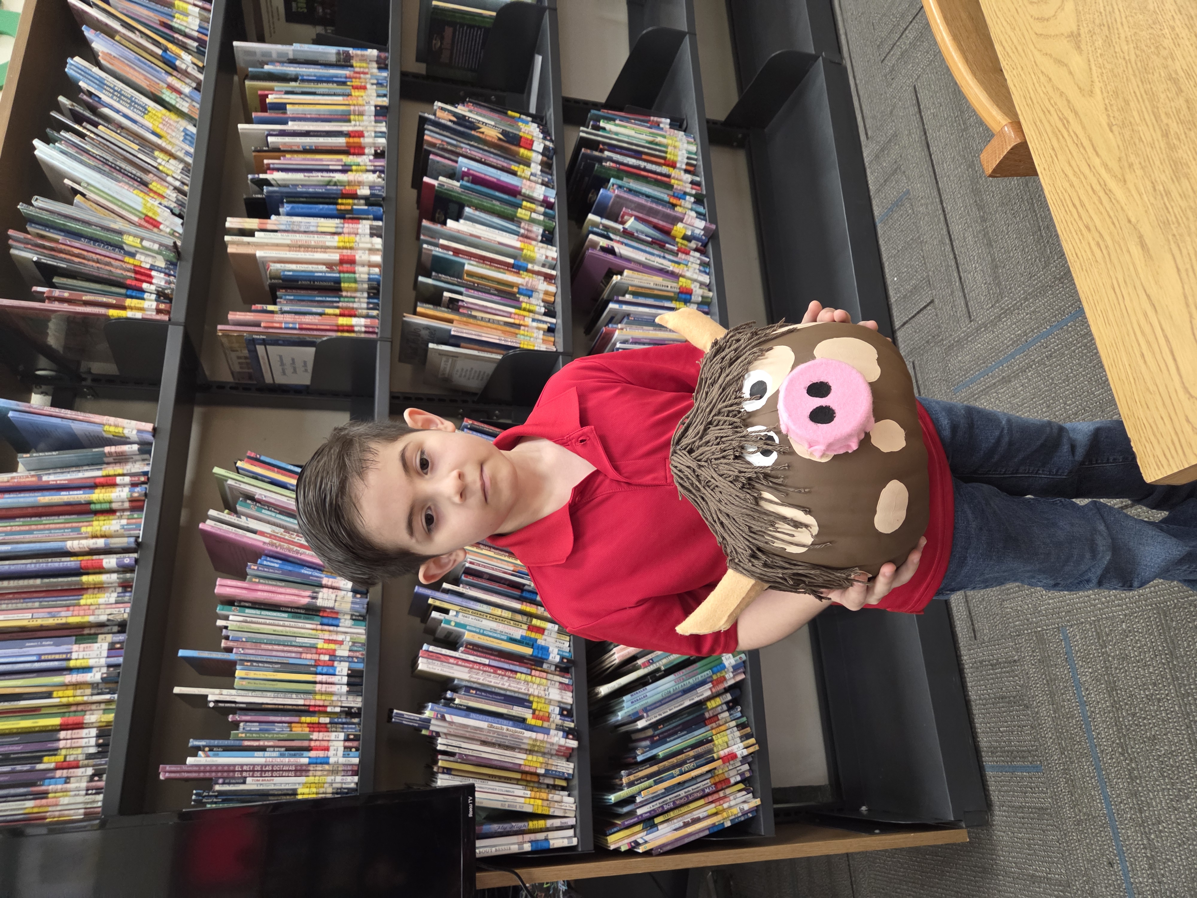 A young boy in a red polo shirt holds a decorated pig craft in a library.
