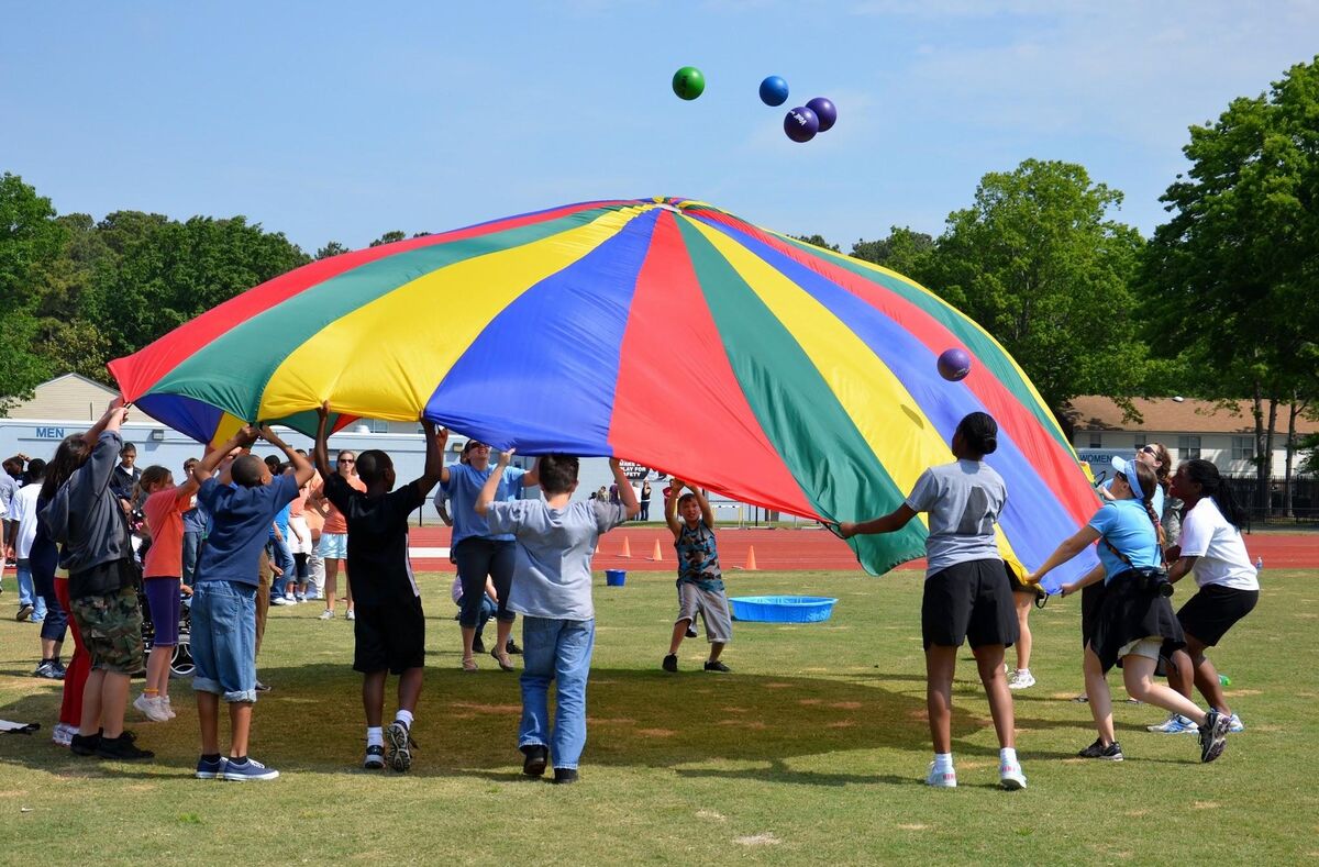 Children and adults hold a large, colorful parachute while playing outdoors on a sunny day.