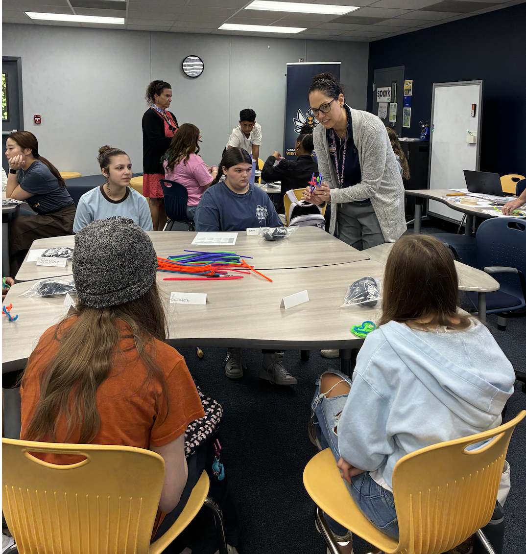 A teacher demonstrates a craft project to a group of students sitting at tables.