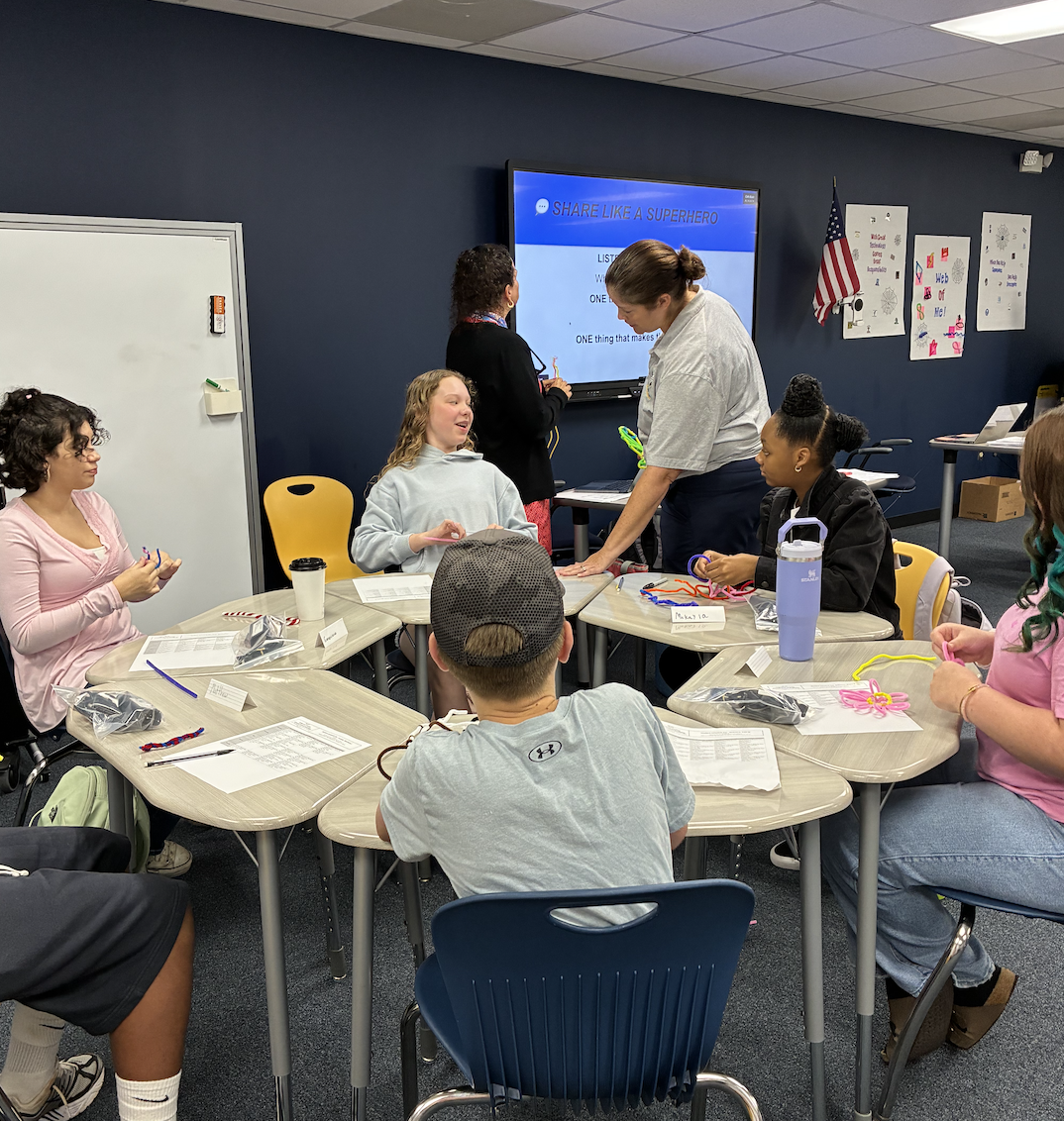Students and teachers engage in a collaborative activity around a clover-shaped table in a classroom.