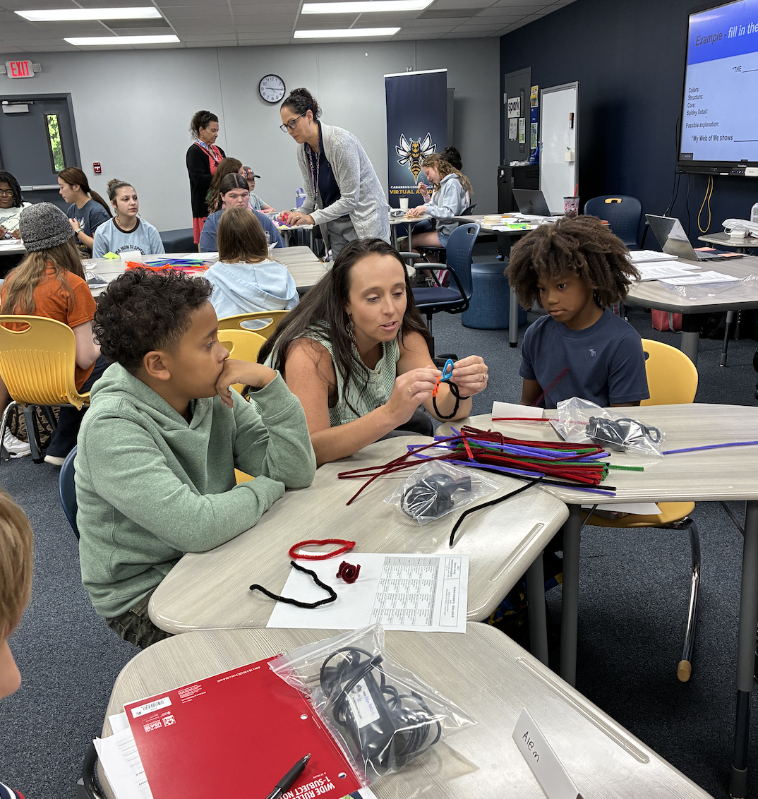 A teacher guides students in a classroom activity, demonstrating with colorful pipe cleaners.