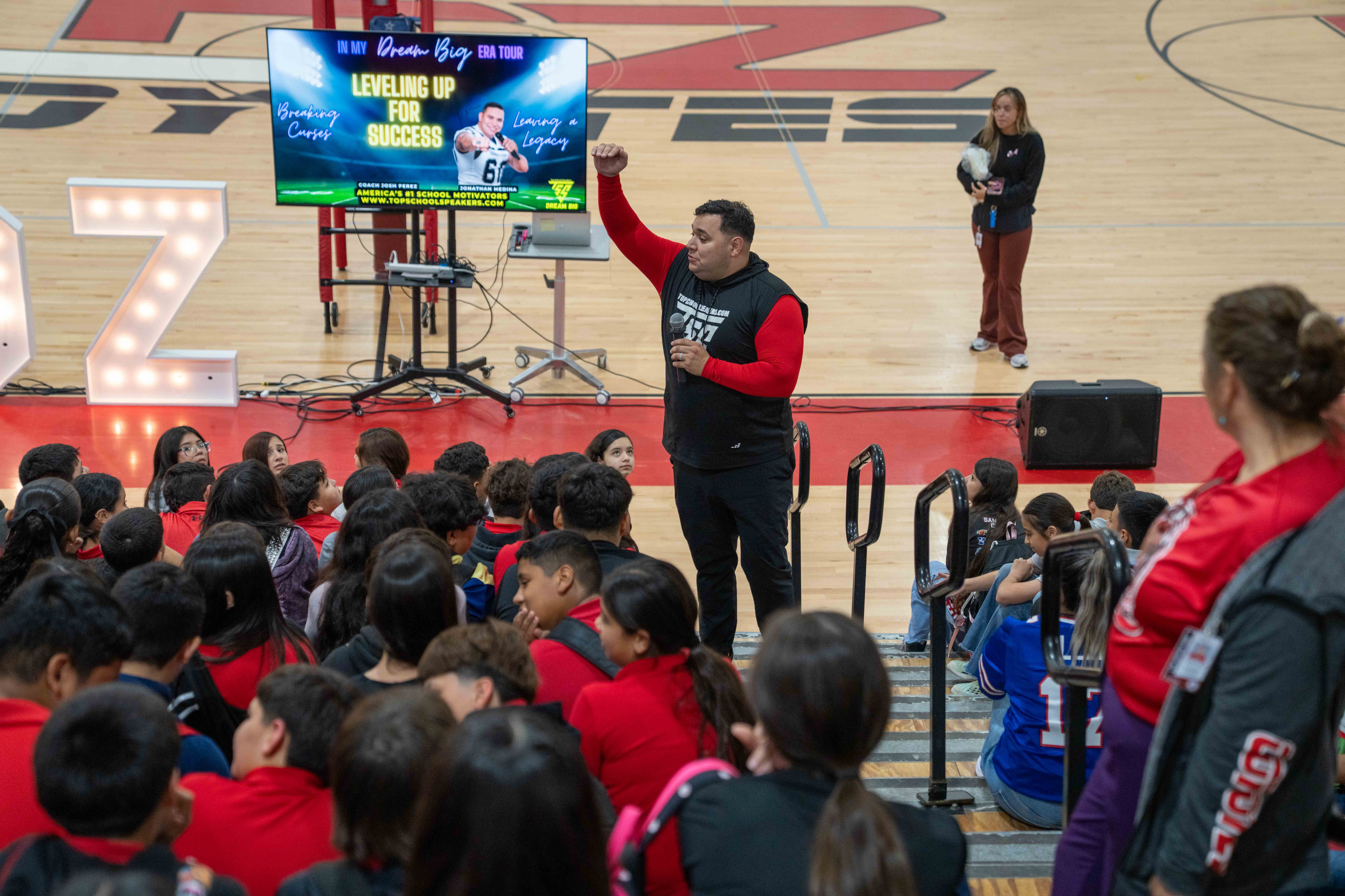 A speaker holds a microphone and raises a fist while addressing a large group of seated students in a gymnasium.