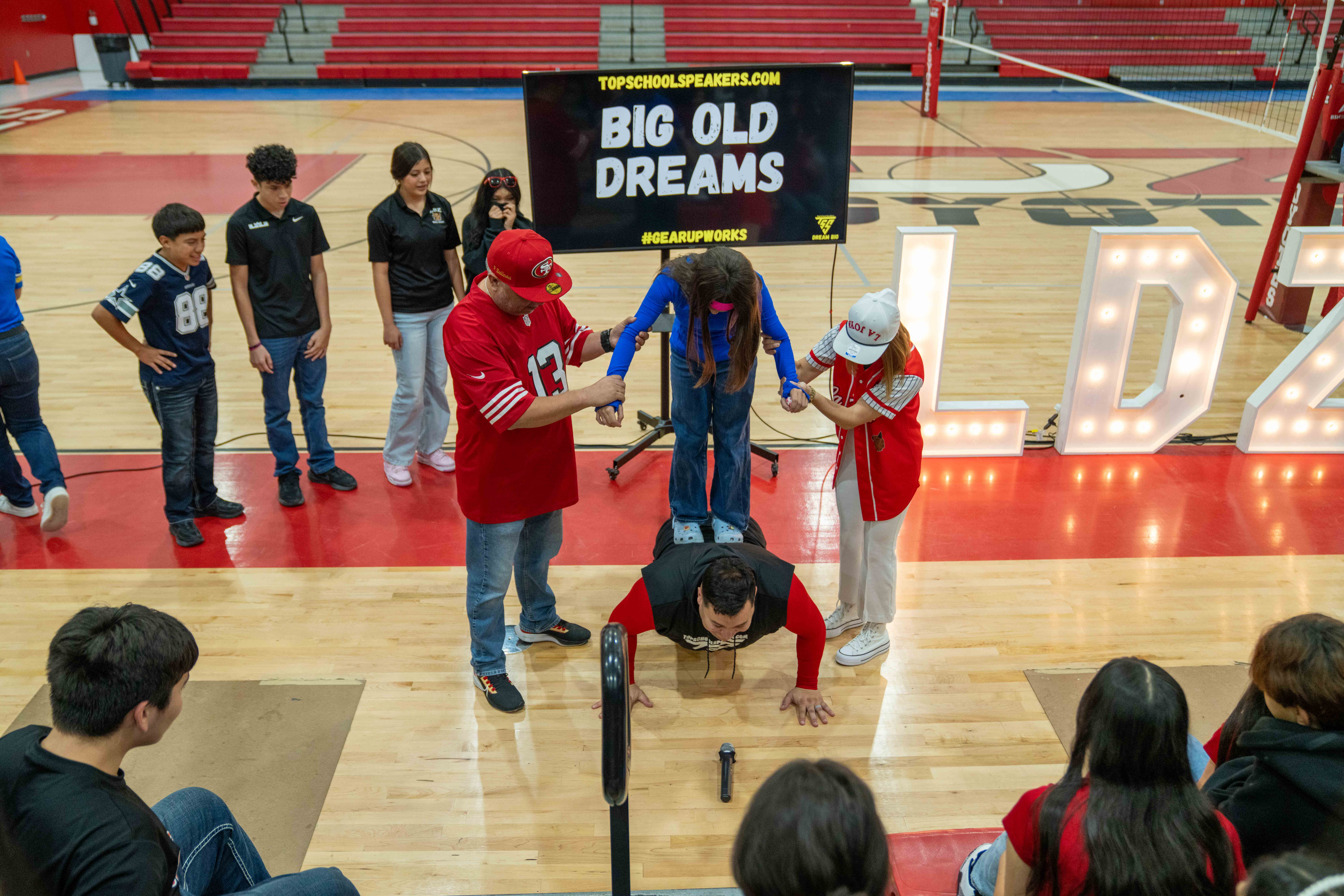 A person does push-ups with a young person standing on their back, assisted by two adults in a school gymnasium.