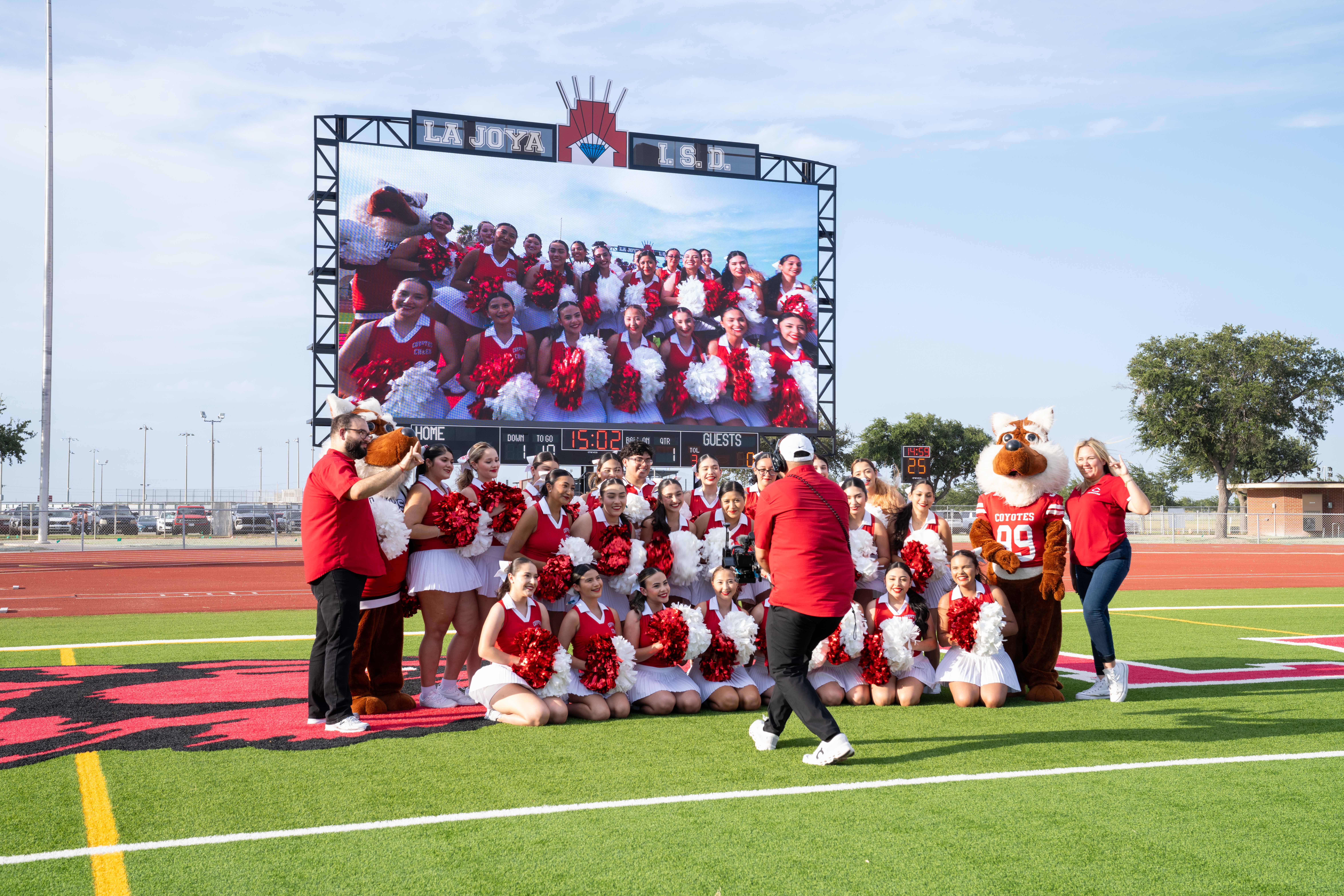 A group of cheerleaders in red and white uniforms pose for a photo on a football field with a mascot.