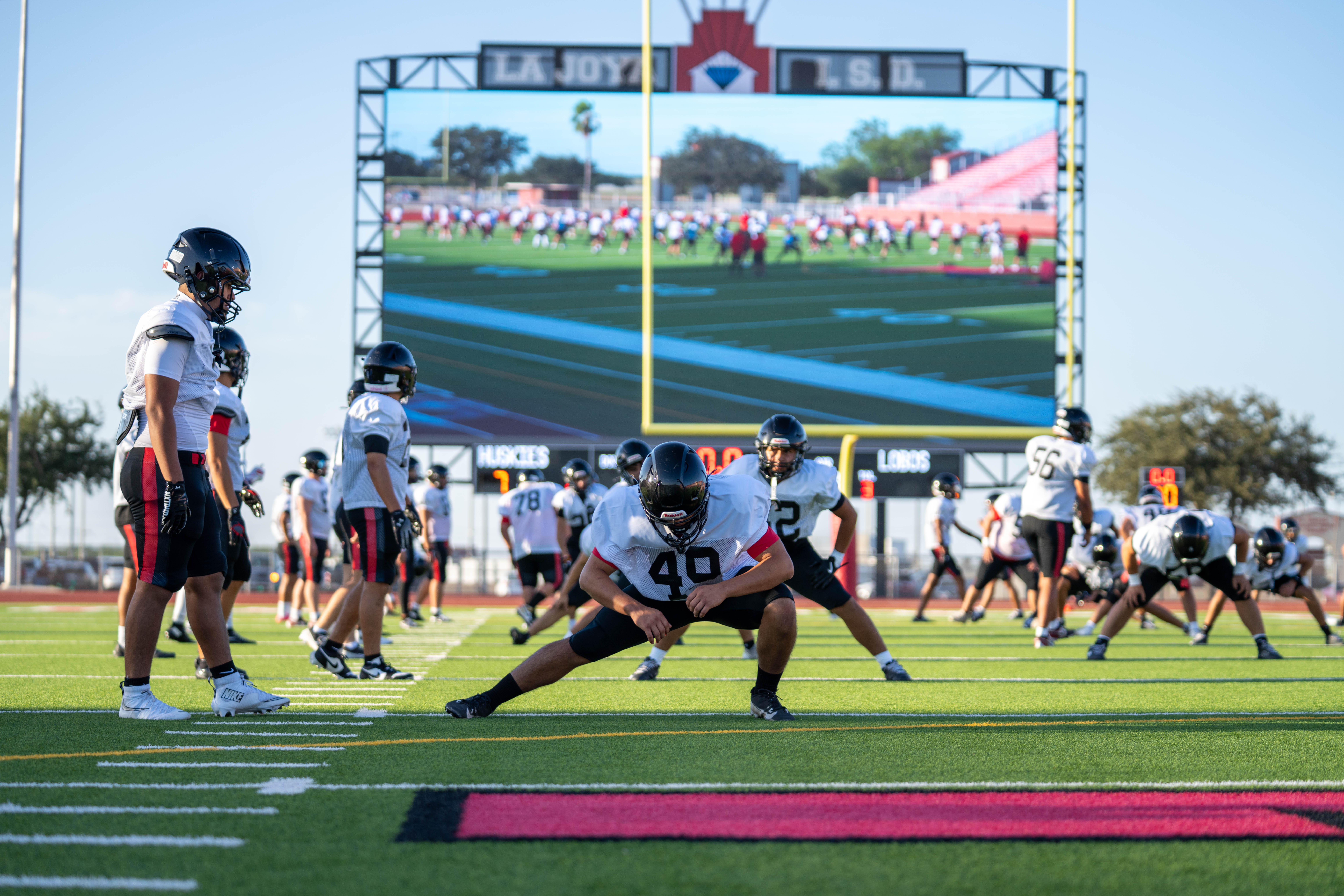 Football players in uniform stretch on a bright green field with a large stadium screen in the background.