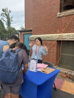 A woman stands behind a blue-covered table, gesturing playfully to people in front of her.