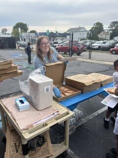 A young woman in a light blue shirt and gloves smiles while holding a pizza box, assisting with a food distribution.